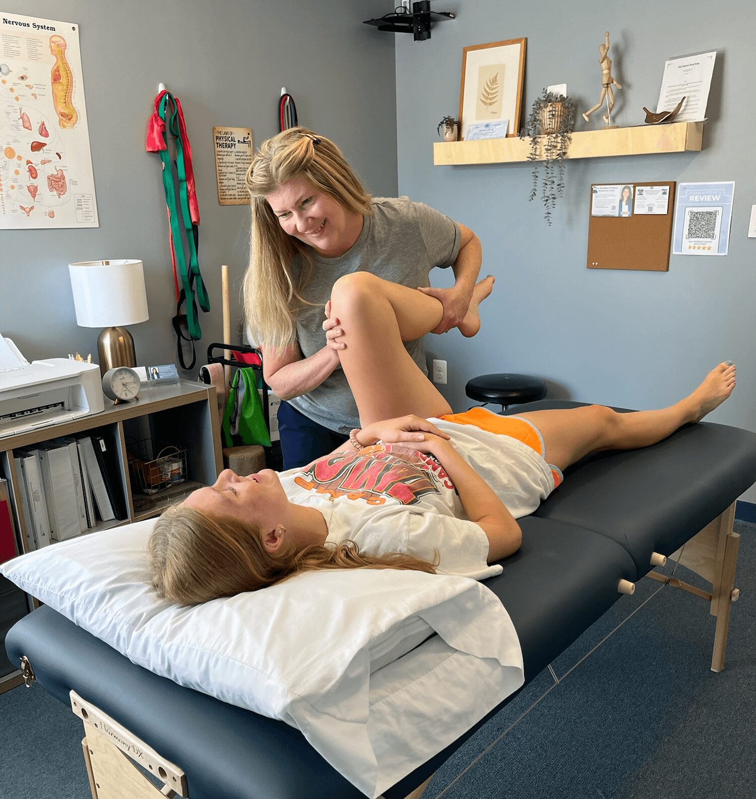 A woman is helping a girl stretch her leg on a medical examination table in a clinic room.
