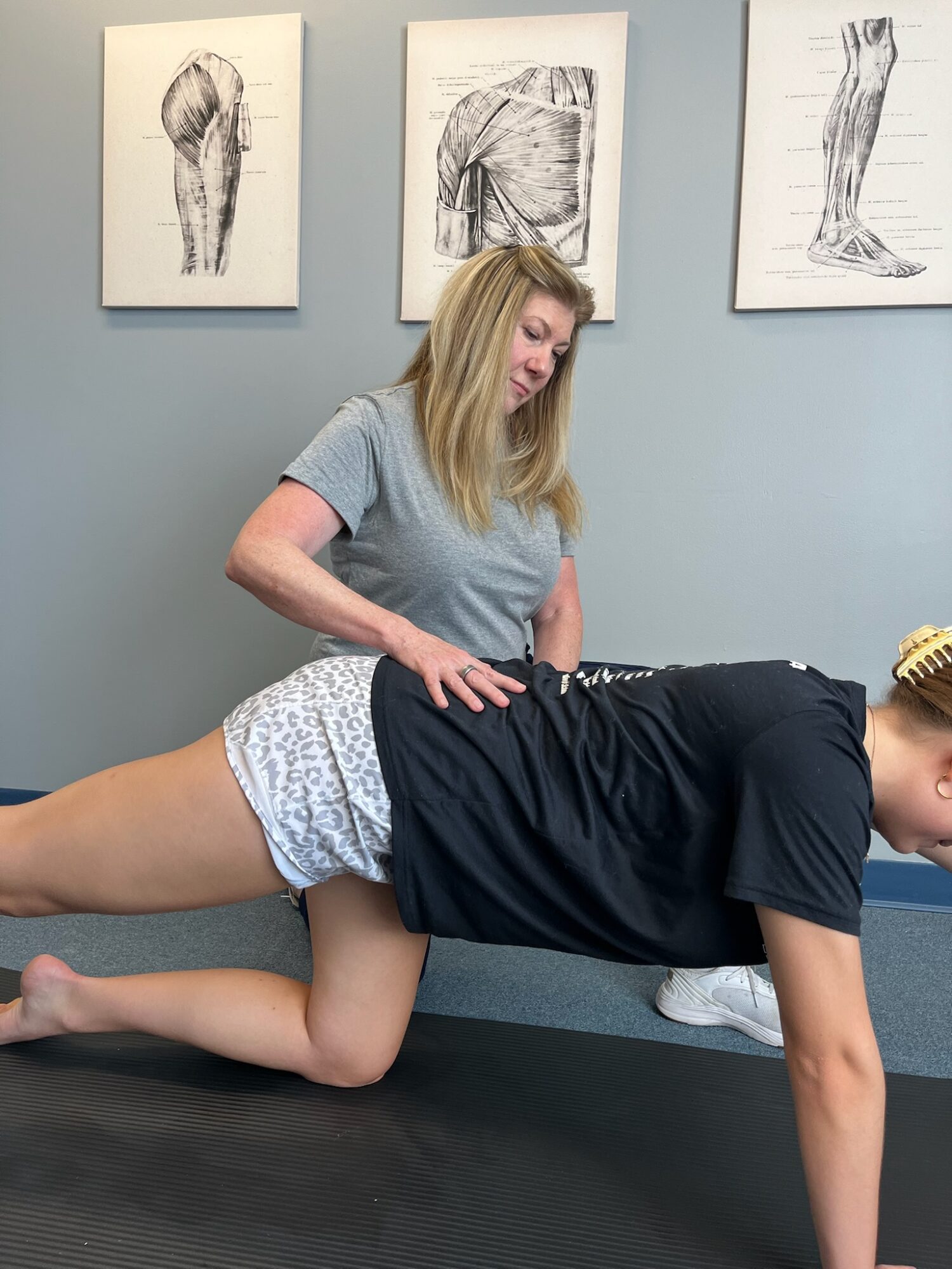 A woman performs a physical therapy exercise on a person in a clinical setting with anatomical drawings on the wall.