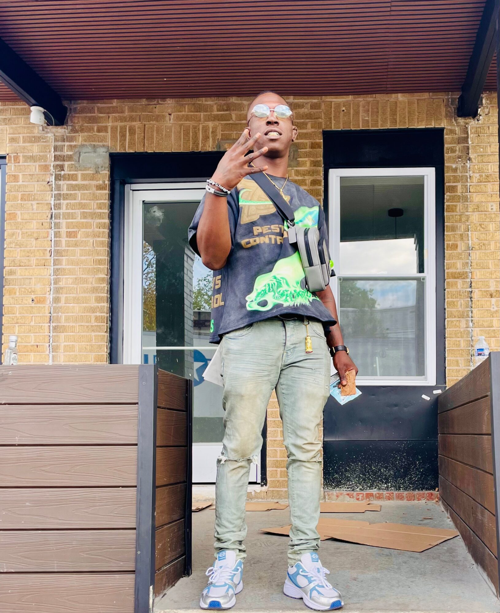 Person standing on a porch with brick walls, wearing sunglasses, a dark t-shirt, light pants, and sneakers, holding a phone.