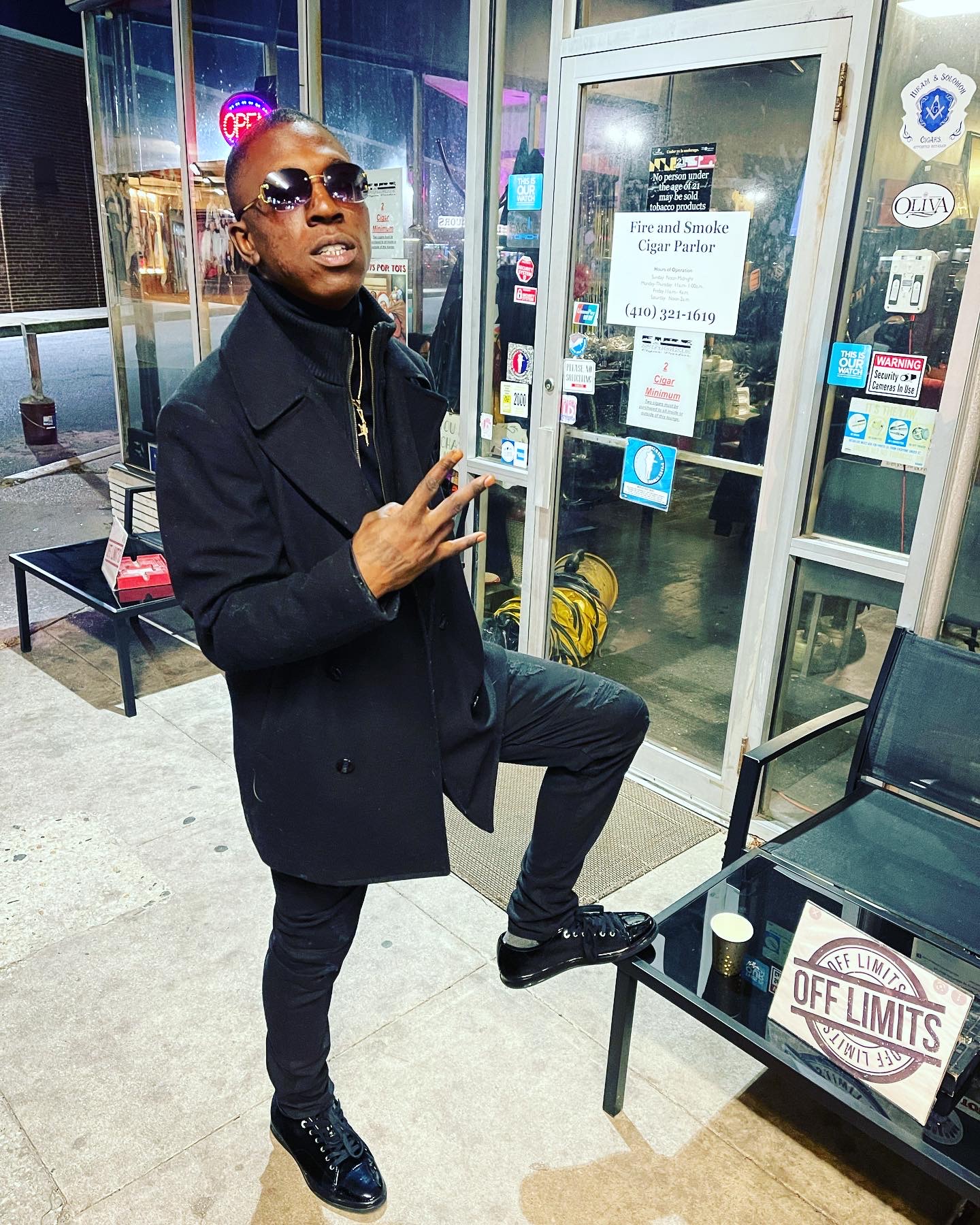 Person with sunglasses making a peace sign pose inside a store near a glass door and bench, with signs and stickers visible.