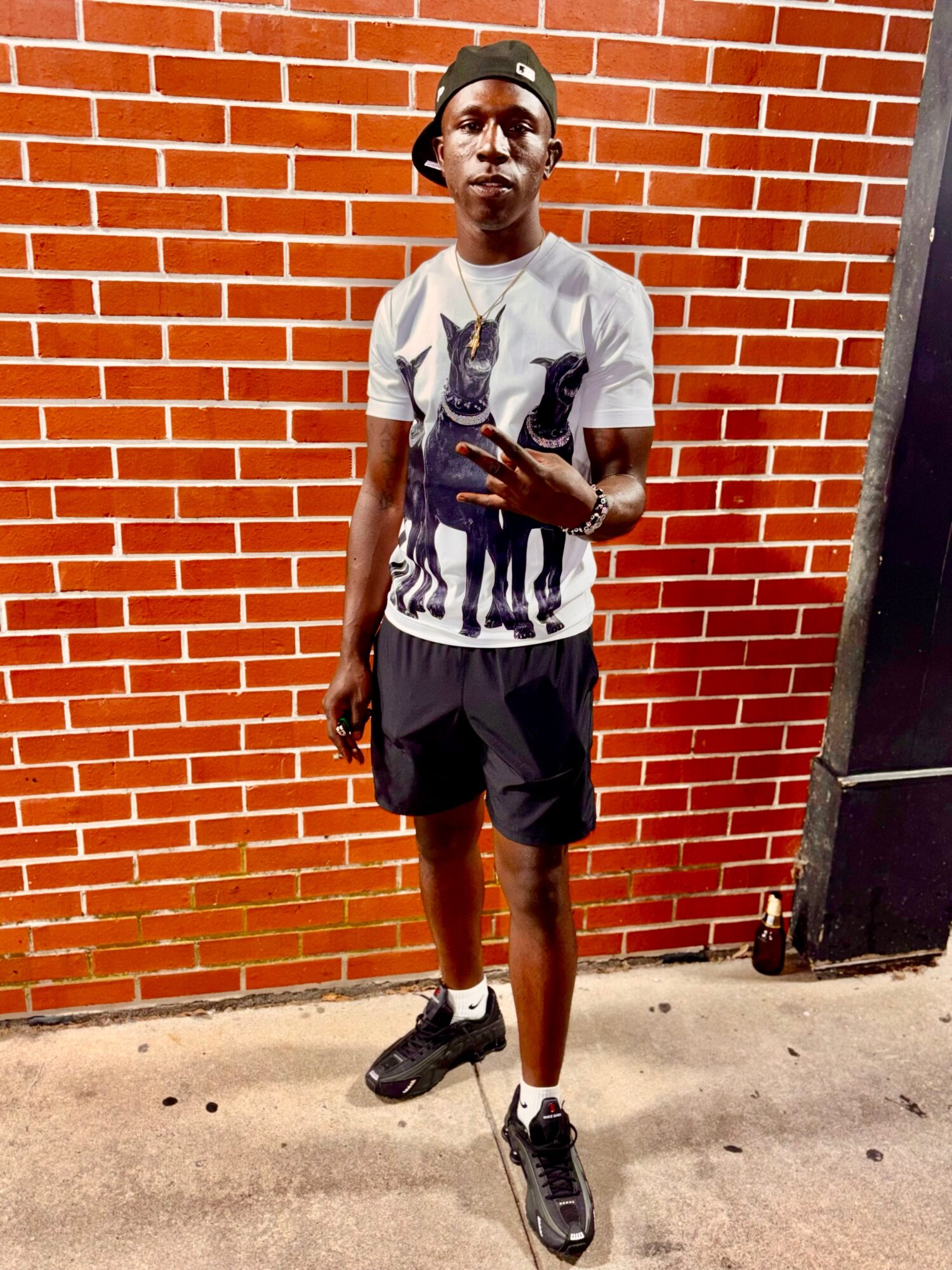 Young man standing against a brick wall, wearing a graphic t-shirt, shorts, and sneakers, holding a phone in one hand.