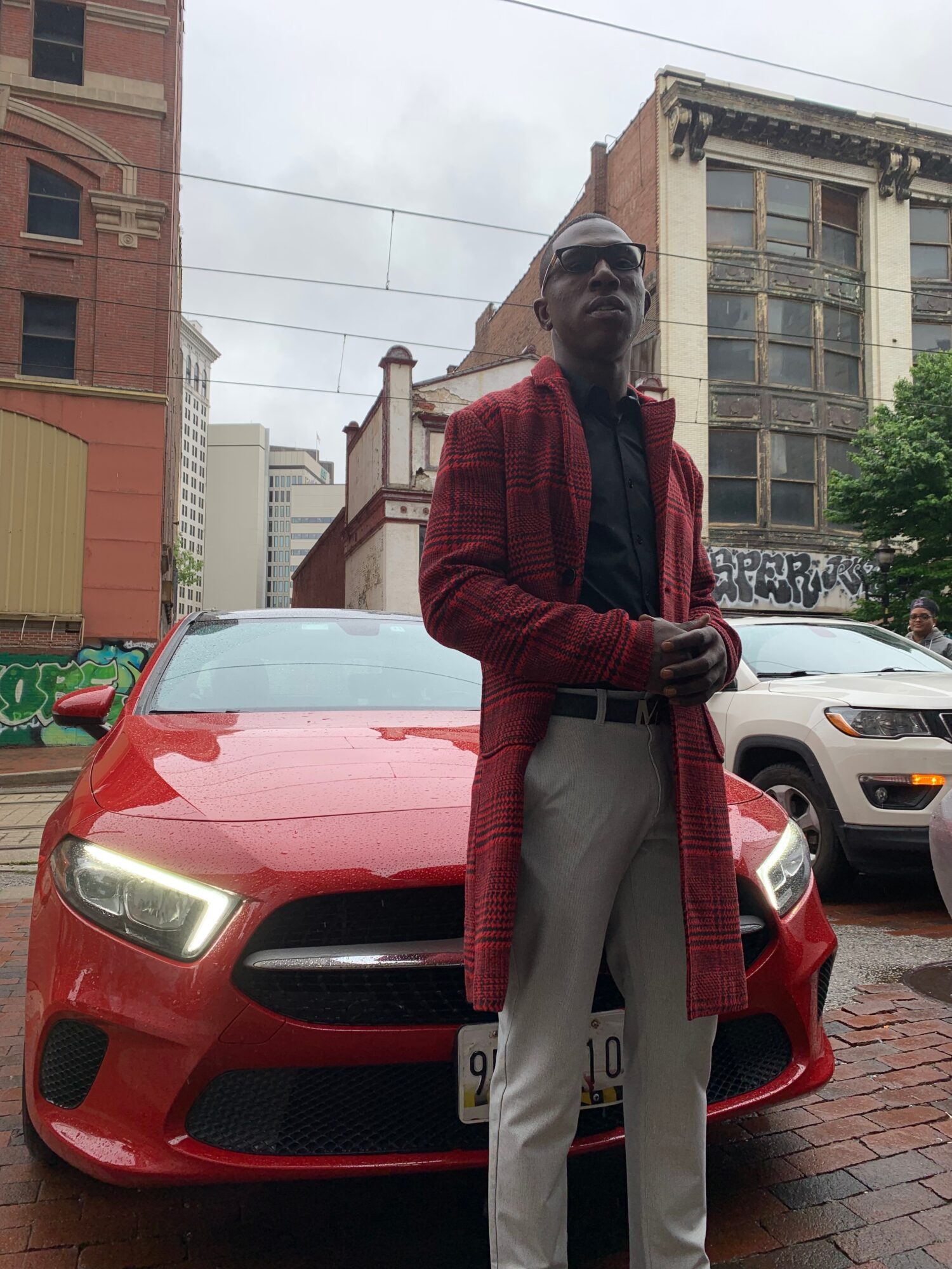 Man in sunglasses wearing a red coat and gray pants standing in front of a red car on a city street.