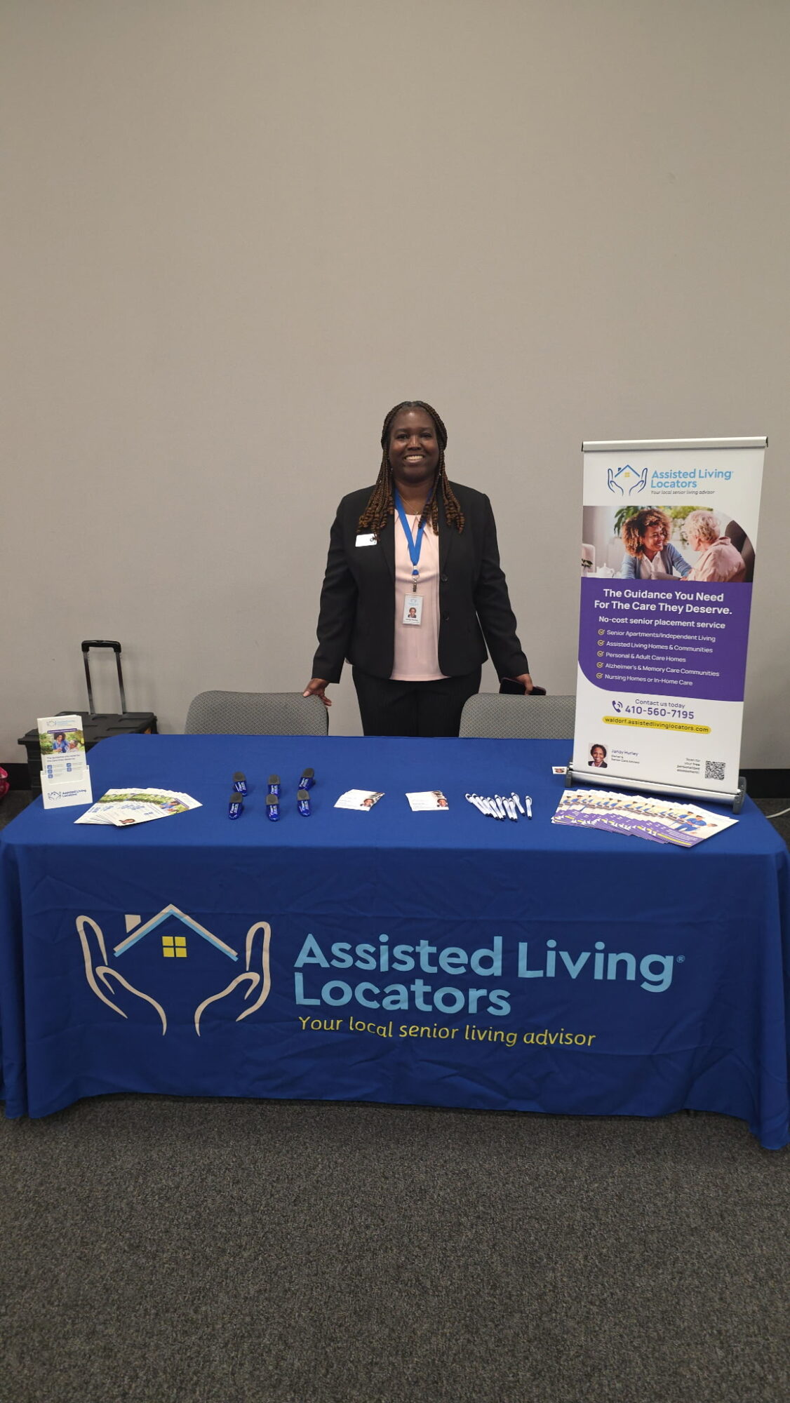 Person standing behind a table with a blue tablecloth displaying the Assisted Living Locators logo, with a poster and brochures nearby.