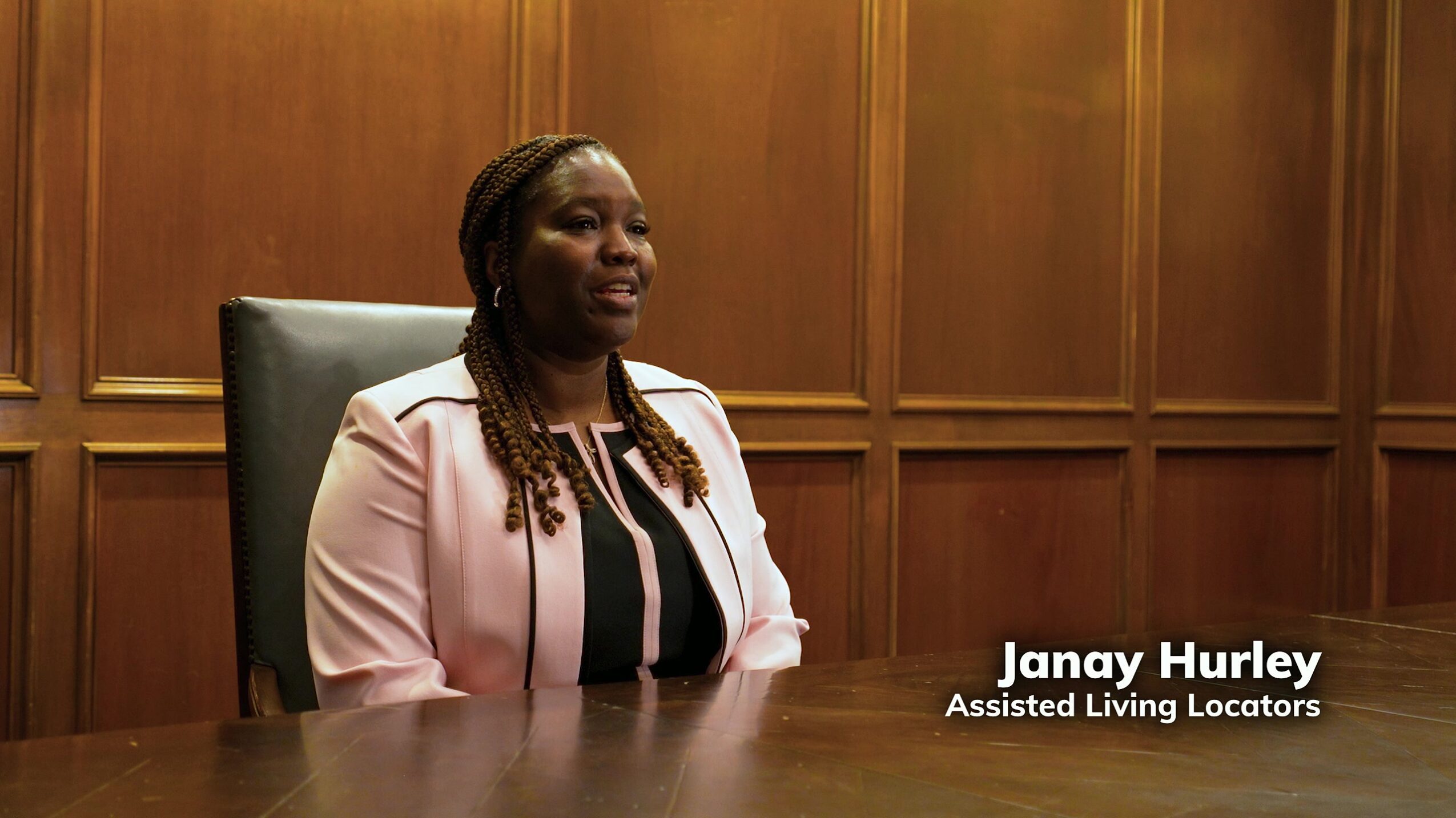 Woman with braided hair sitting at a table in a wood-paneled room, wearing a light pink blazer and black top.