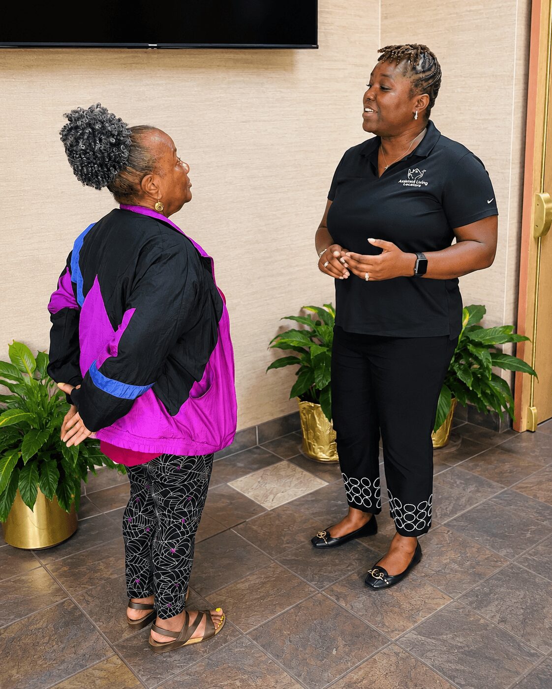 Two women are talking indoors, one wearing a black and purple jacket and the other in a black uniform, with plants behind them.