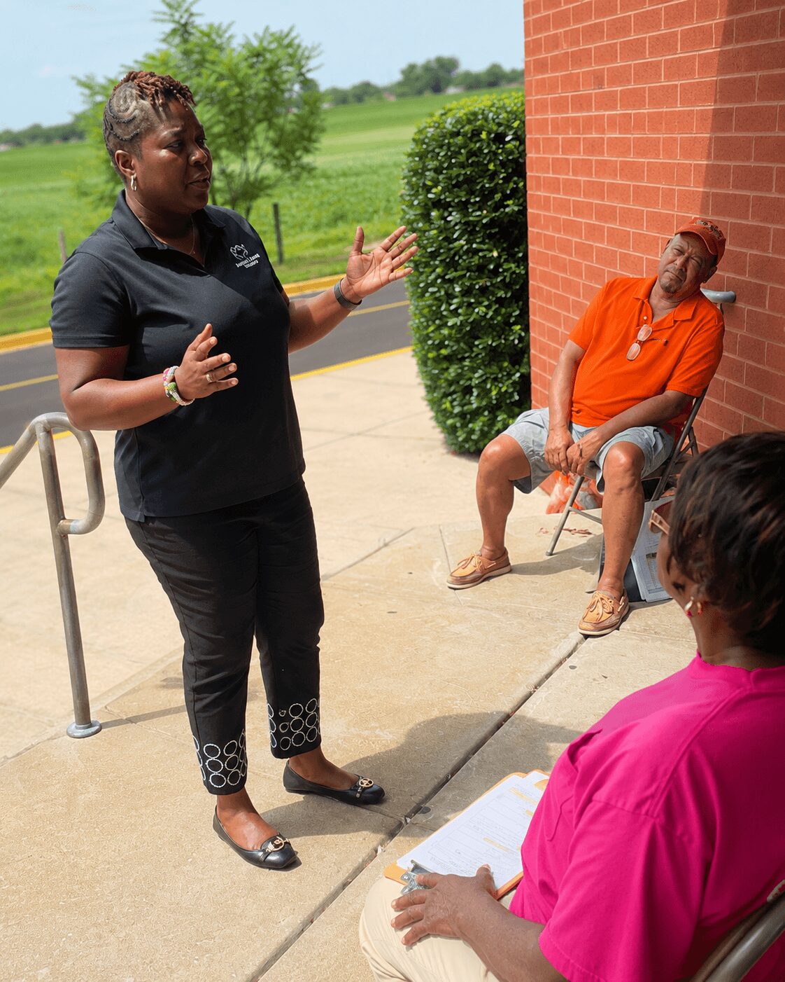 Woman in black speaking outdoors, two women seated nearby, one in orange and one in pink, on a sidewalk with greenery and a brick wall.