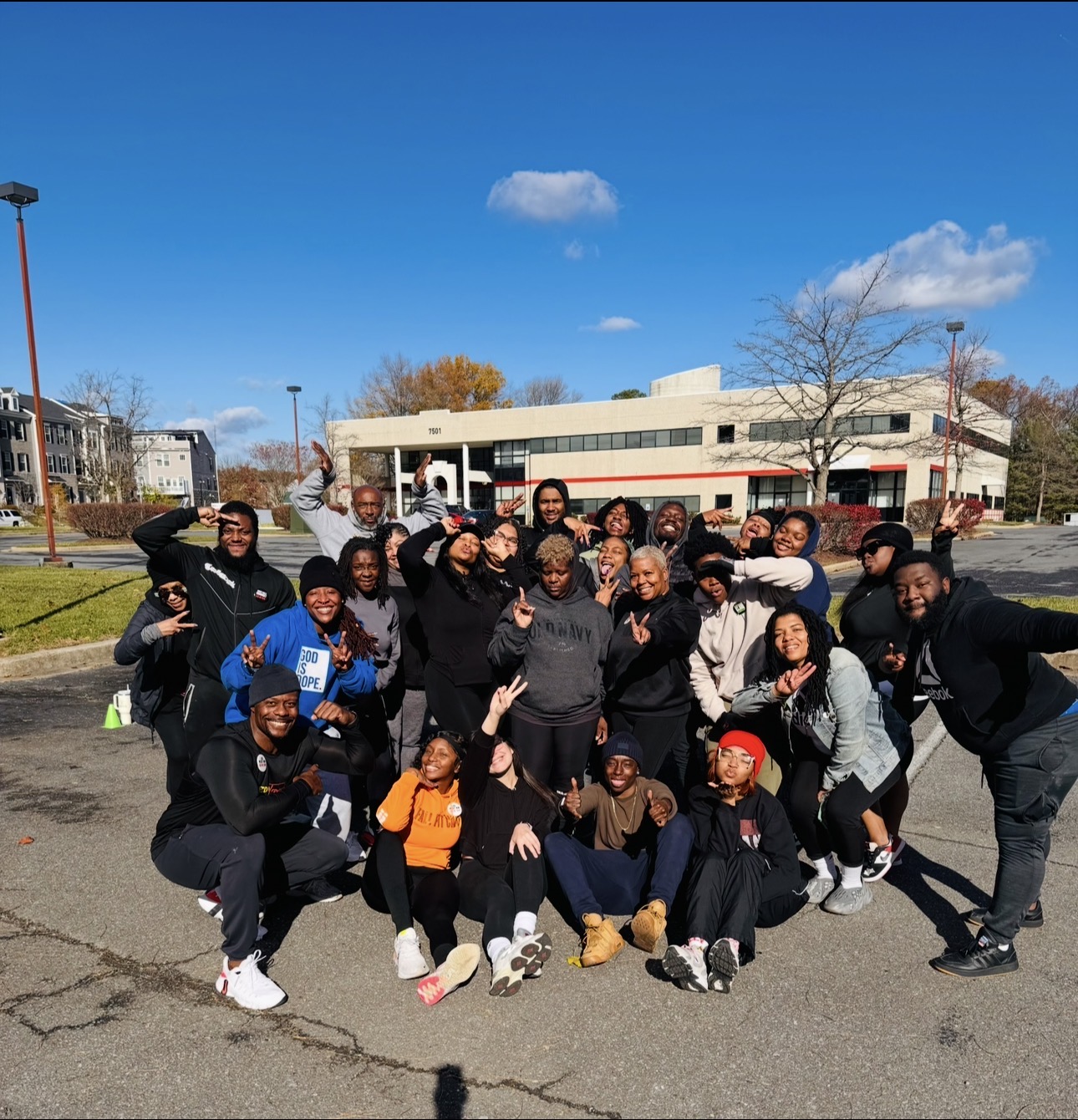 Group of people outdoors in a parking lot, smiling and posing for a photo with a building and trees in the background.