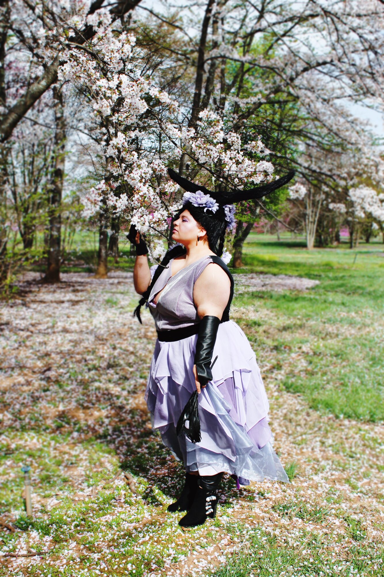 Person in gothic attire standing under blooming cherry blossom tree outdoors, holding a flower, with grass and trees in background.