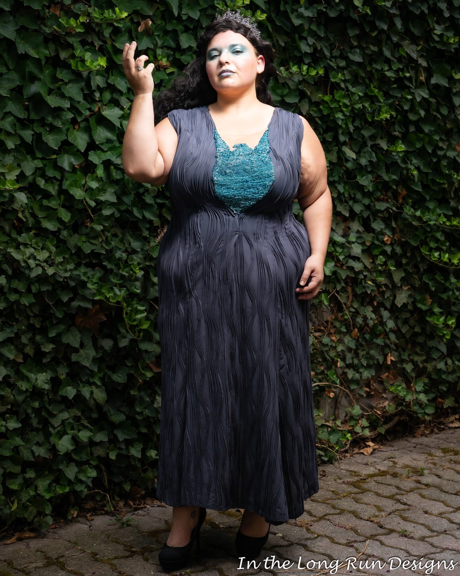 A woman with dark curly hair stands outdoors in front of green foliage, wearing a long black dress with teal details, holding a cigarette, with one hand raised.