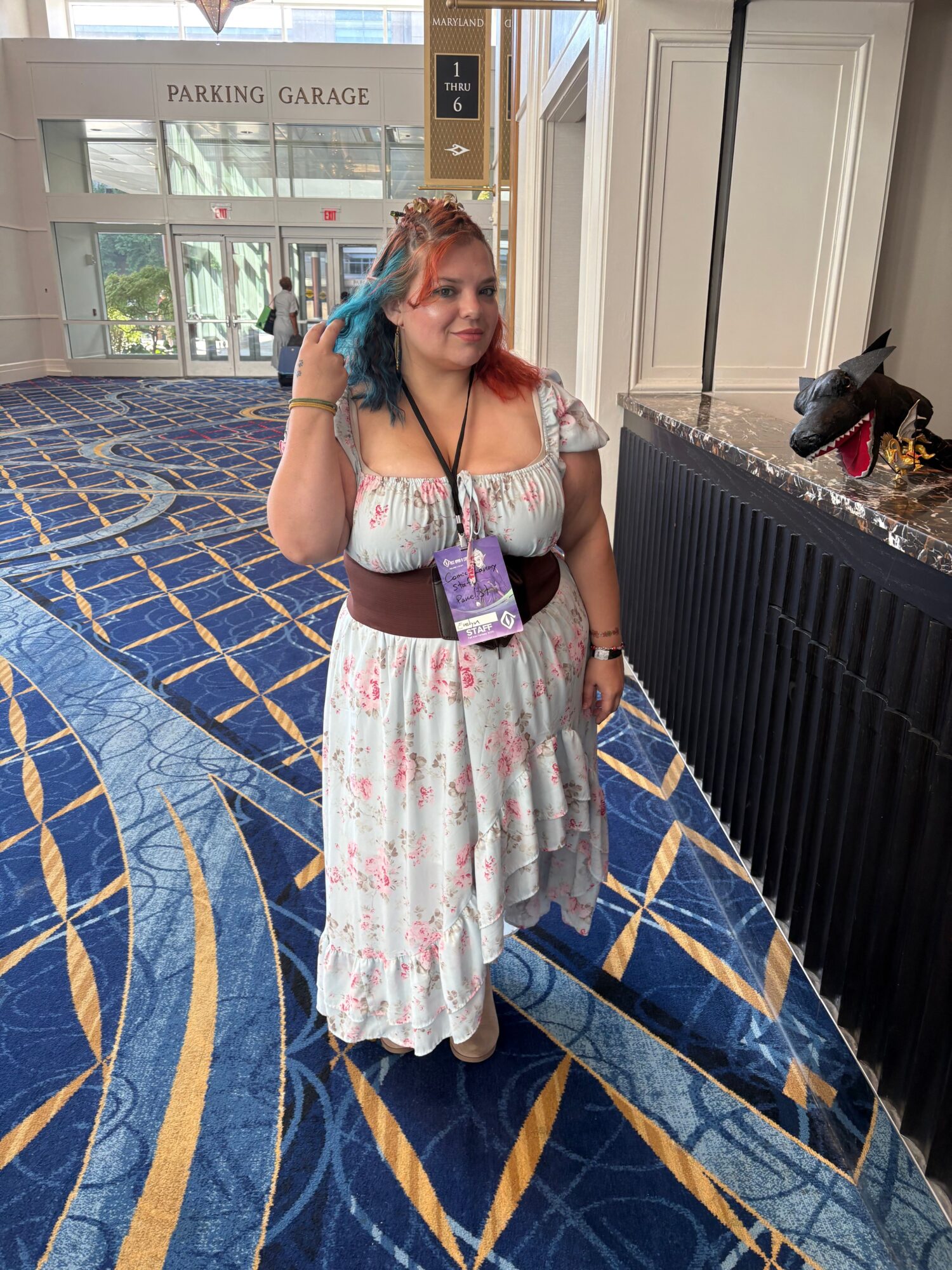 Woman in a floral dress holding a purple bag, standing in a hotel hallway with a patterned carpet and white doors.