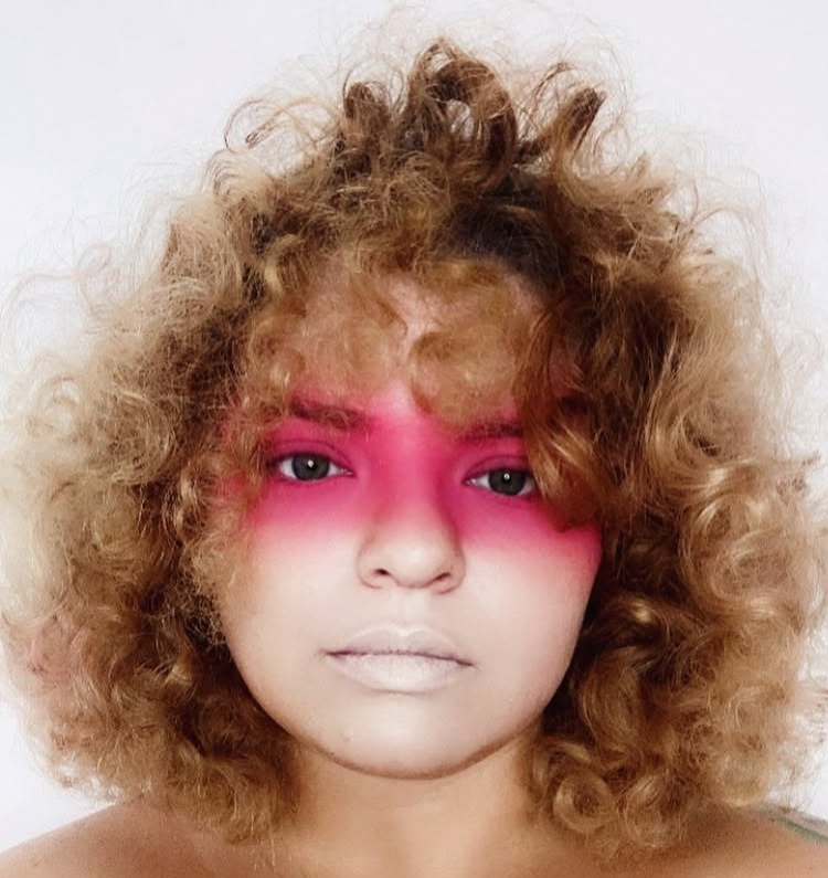 Person with curly hair and pink makeup around eyes, lips painted white, facing forward.