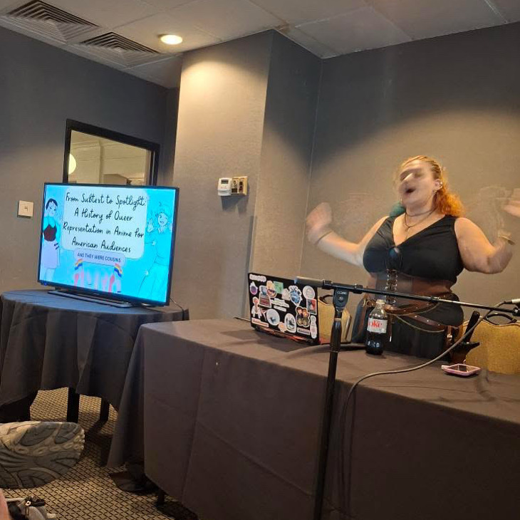 Woman speaking at a table with a laptop and microphone, a TV screen displays a presentation slide, in a room with gray walls.