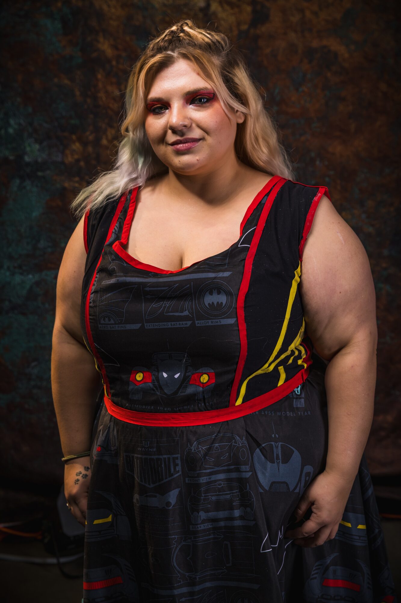 Woman with blonde and pink hair wearing a black and red dress with a racing car design, standing against a dark background.