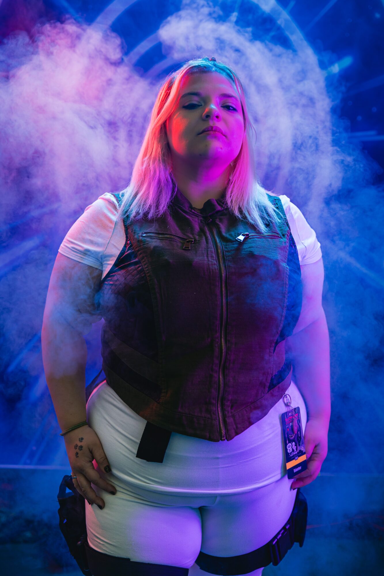 Young woman with colorful hair stands in a smoky, vibrant setting, wearing a vest and white pants, looking confidently at the camera.
