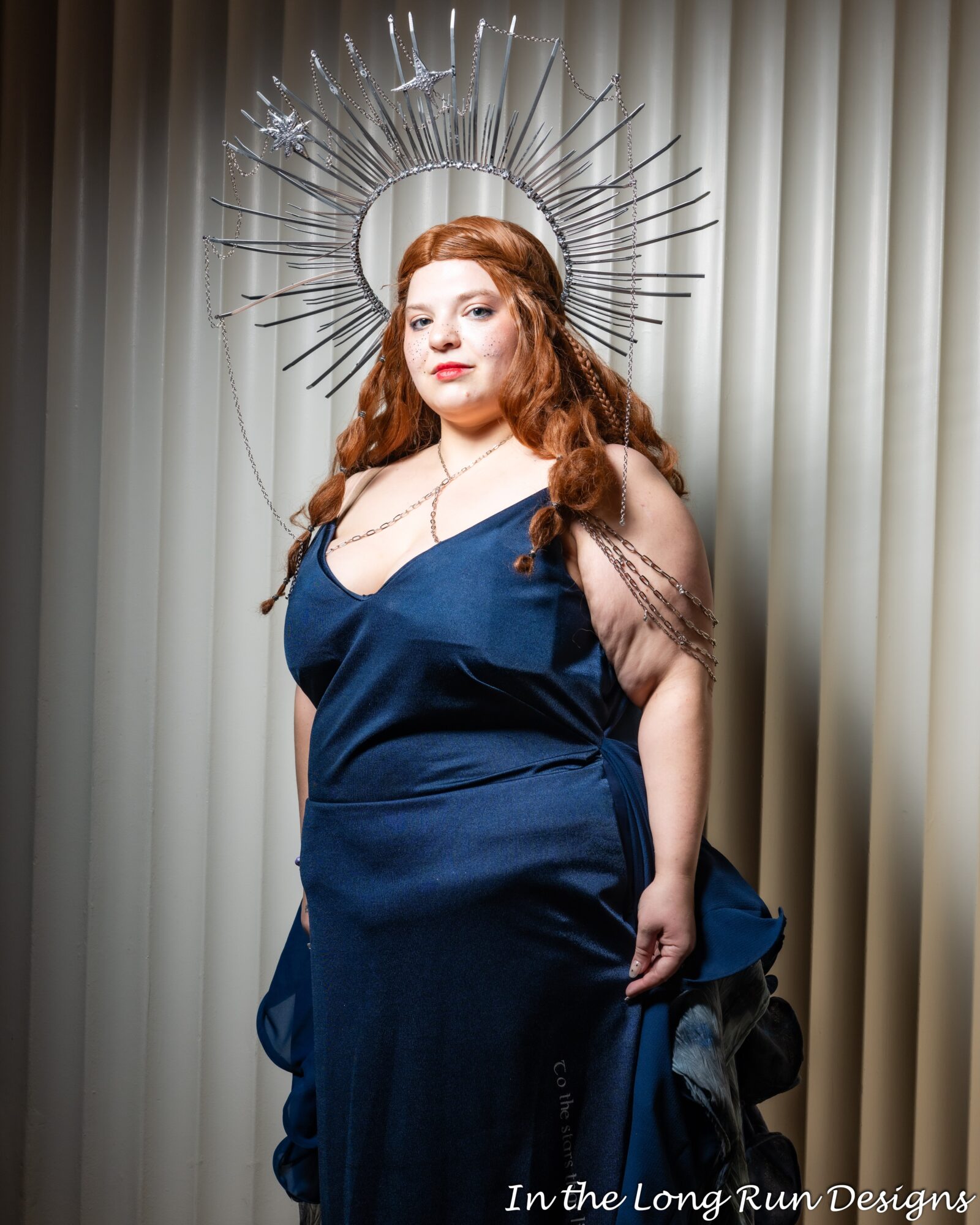 Woman with long red hair in a blue dress, standing in front of curtains, wearing a halo-like headpiece, looking at camera.