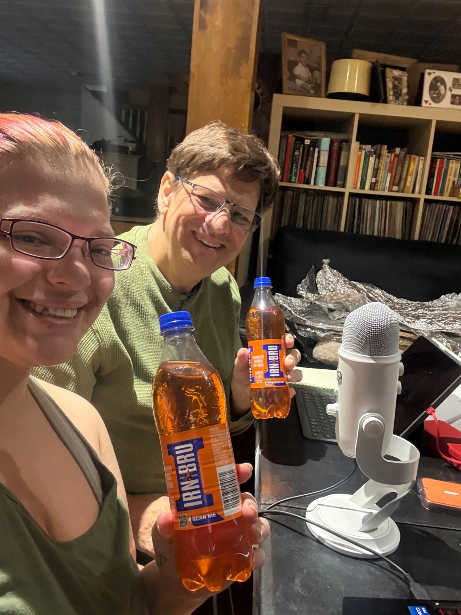 Two people smiling, one holding a bottle of IRN-BRU soda, in a room with bookshelves and a table with various items.