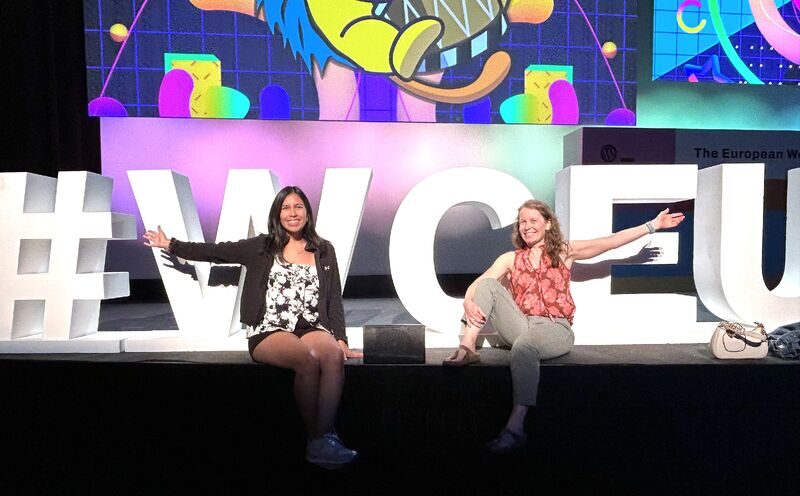 Two women sit on stage in front of large white hashtag signs, smiling and gesturing with arms outstretched, with colorful cartoon background.