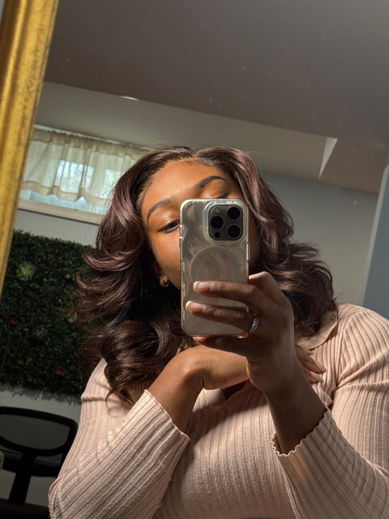 Person with wavy hair taking a mirror selfie with a smartphone, wearing a light-colored sweater, in a room with a window and greenery outside.