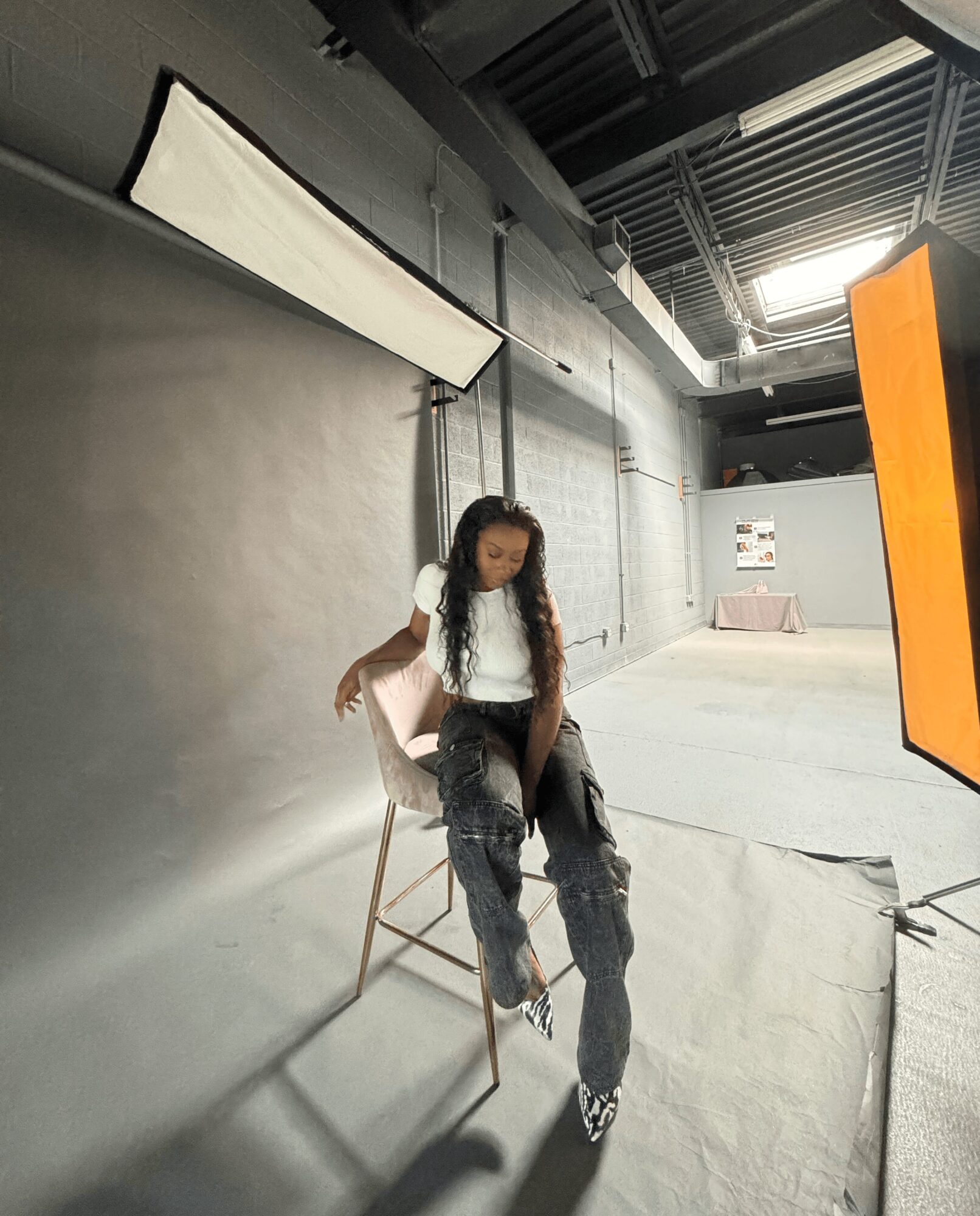 Young woman sitting on a chair in a photography studio with gray walls and equipment, looking down, with studio lights overhead.
