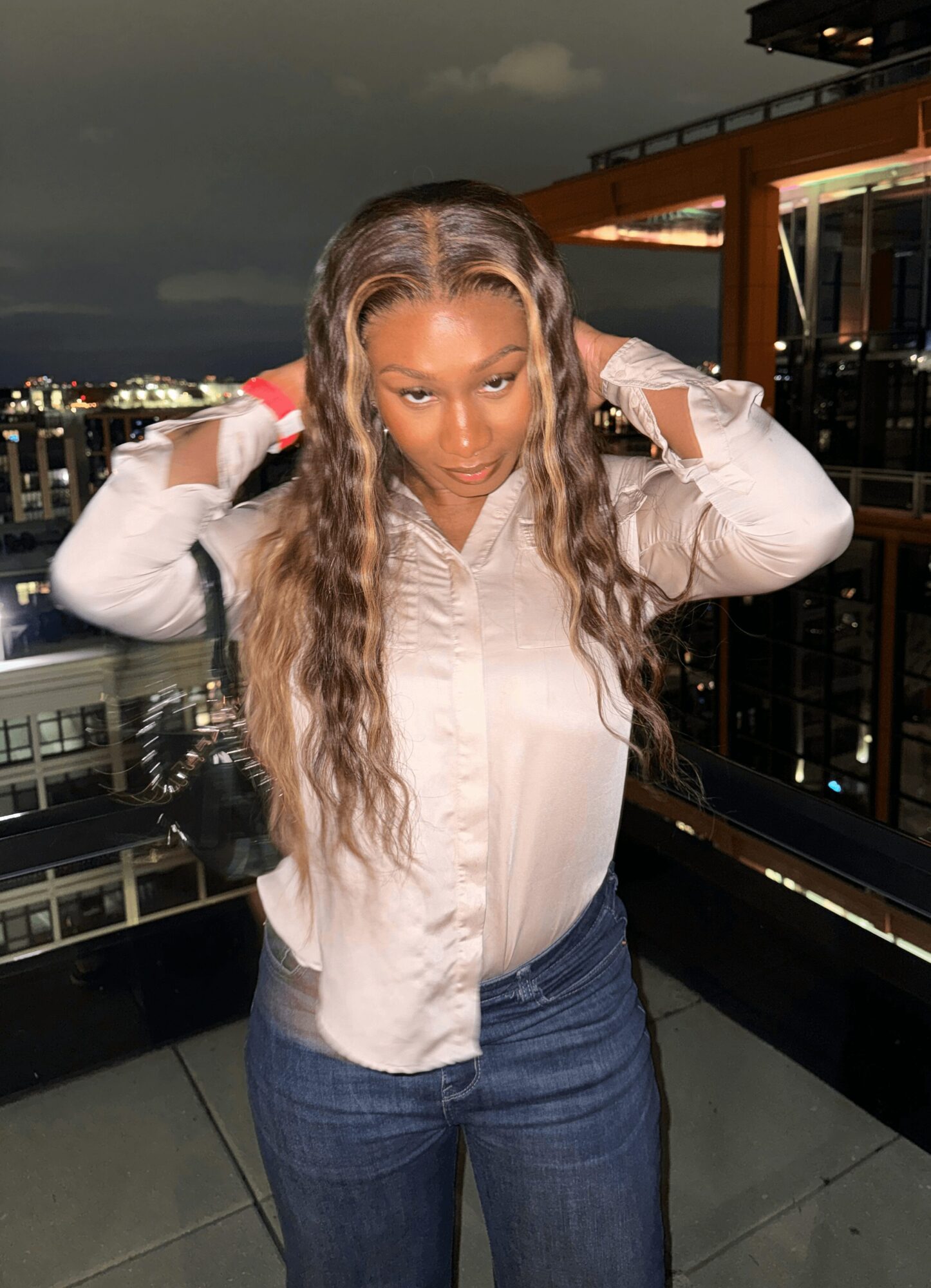 Woman with long wavy hair adjusting her hair on a rooftop at night, city lights in background.