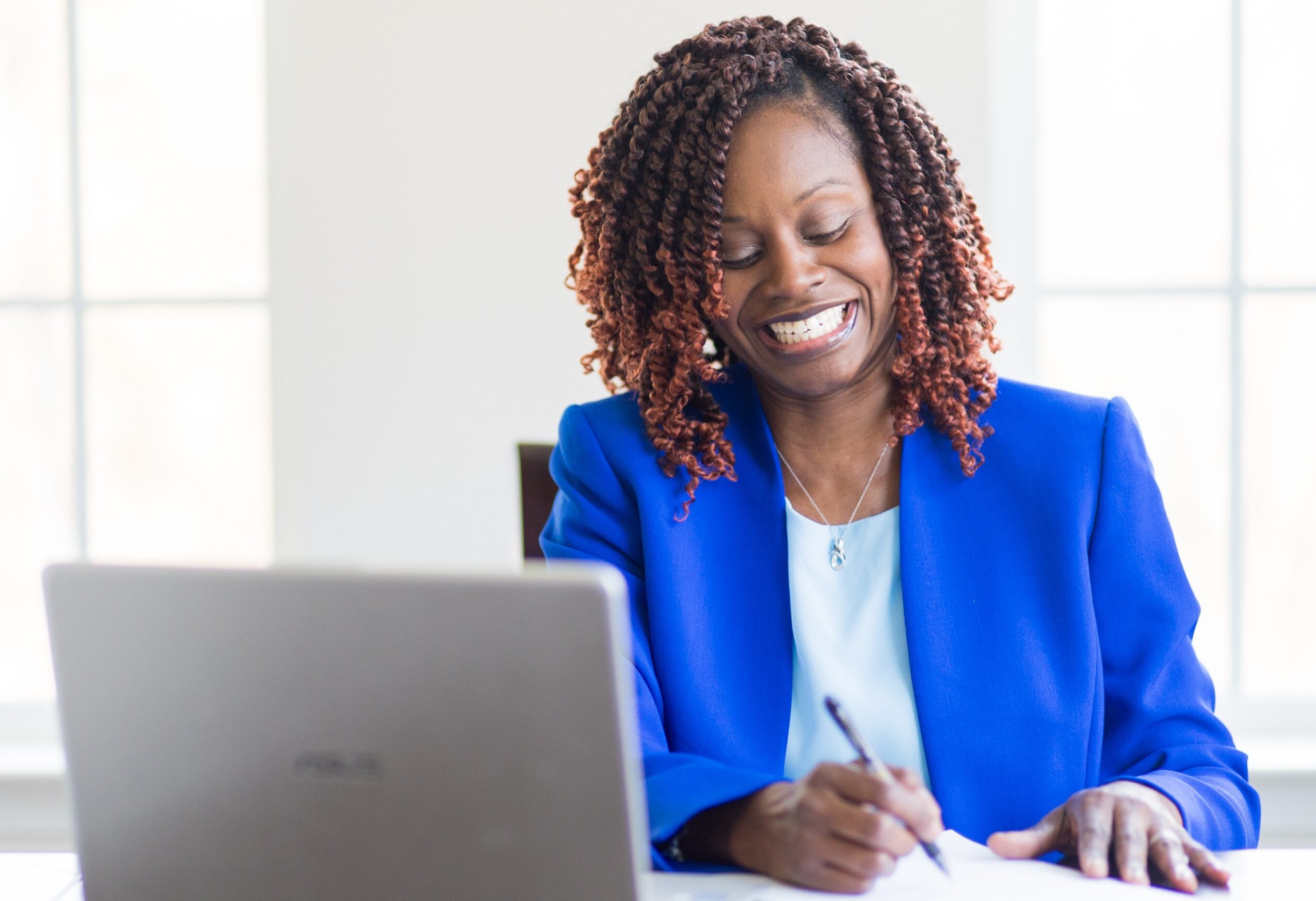 Woman with curly hair smiling, wearing a blue blazer, sitting at a desk with a laptop, writing on paper.