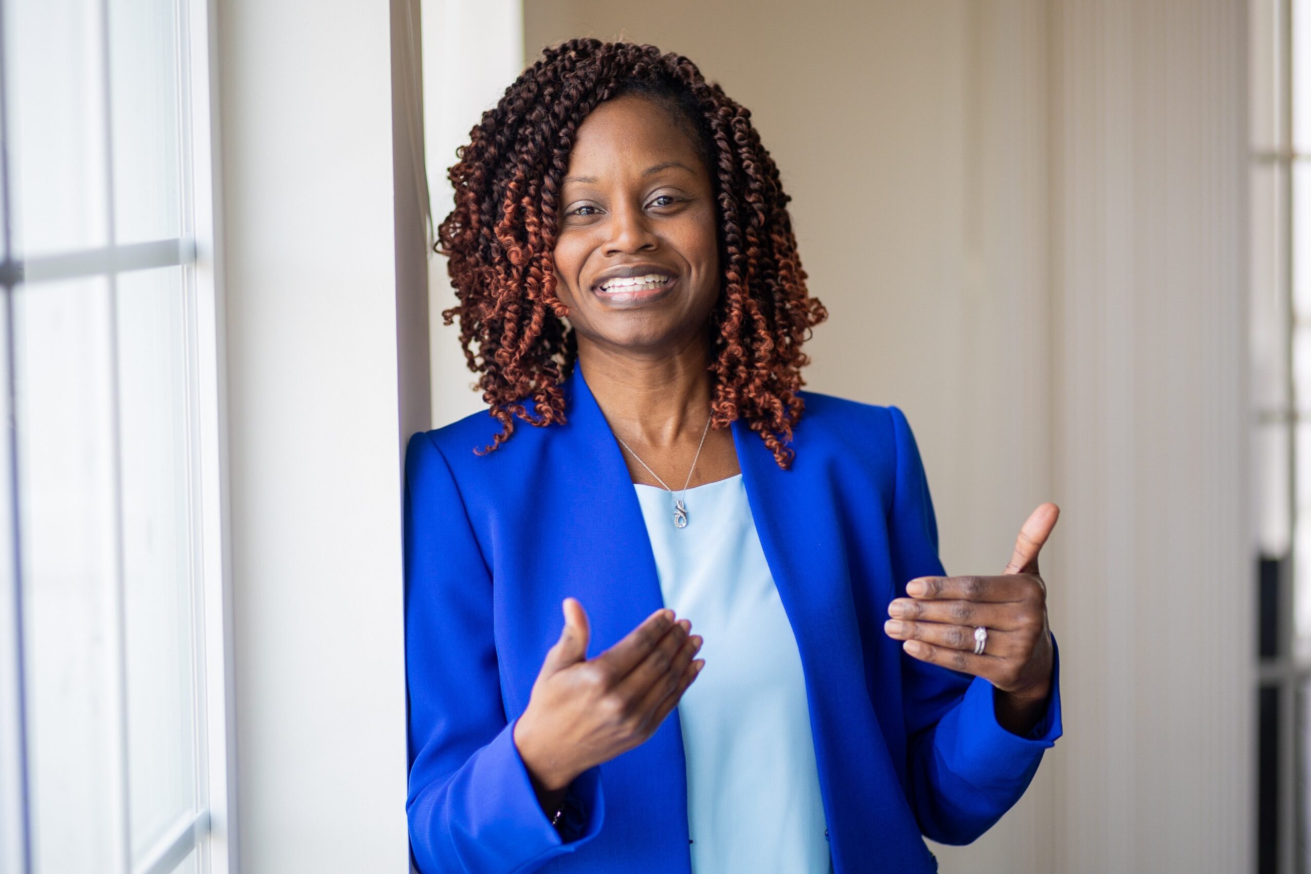 Woman with curly hair wearing a blue blazer, smiling and gesturing with hands indoors.