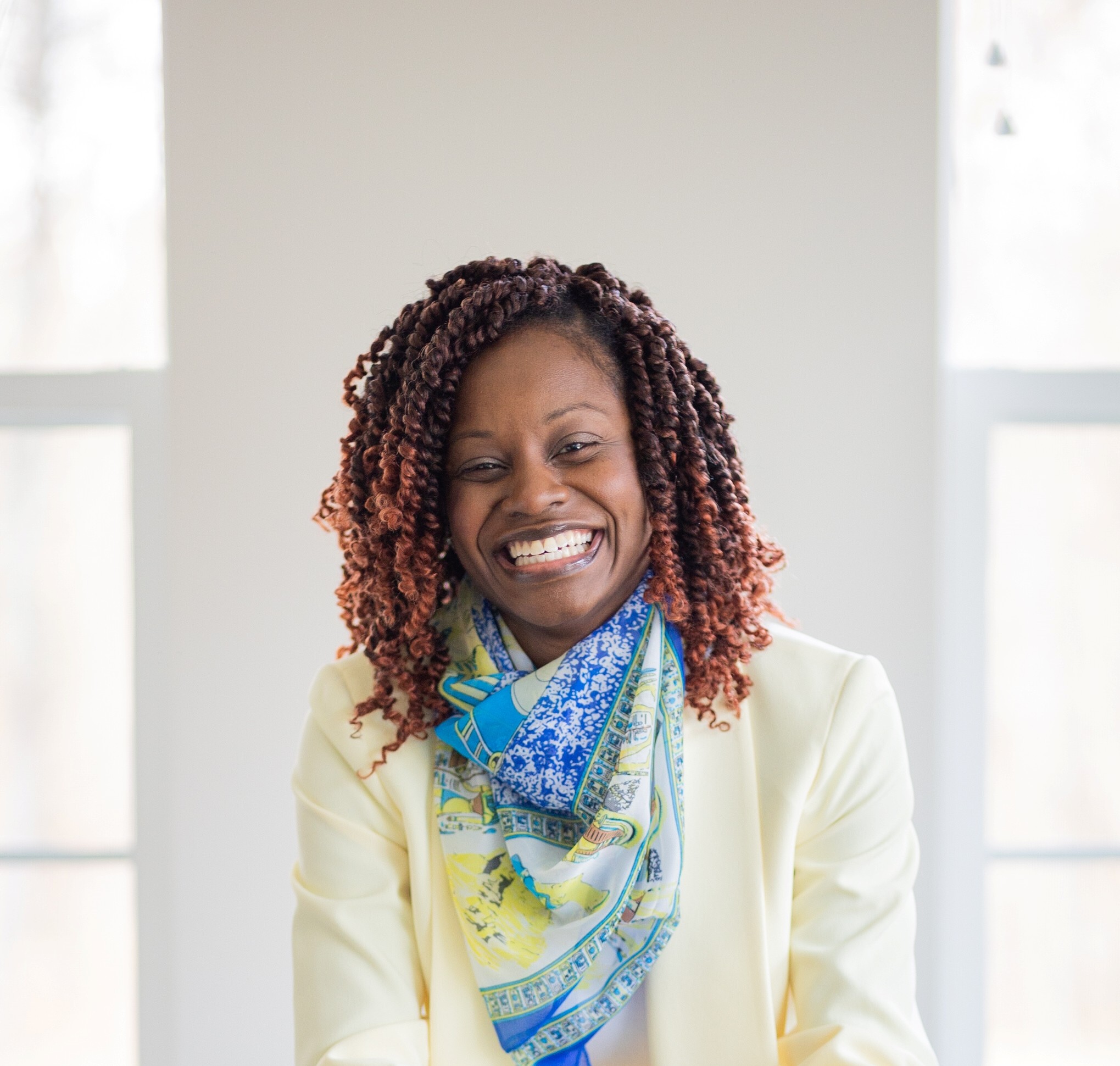 Woman with curly hair smiling, wearing a light-colored blazer and a colorful scarf, standing indoors with windows behind her.