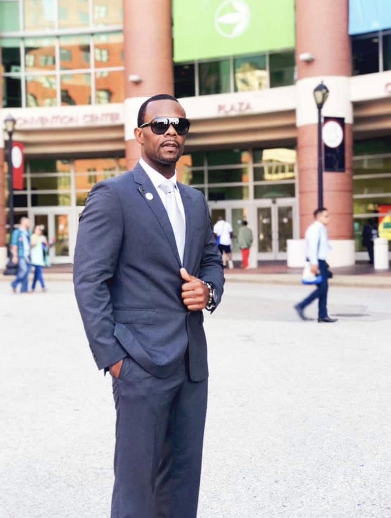 Man in a suit and sunglasses standing outdoors in an urban area with people and buildings in the background.