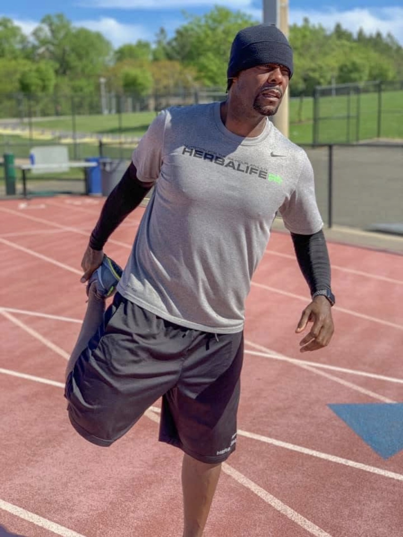Man in athletic clothing stretching on a running track outdoors, with trees and a fence in the background.