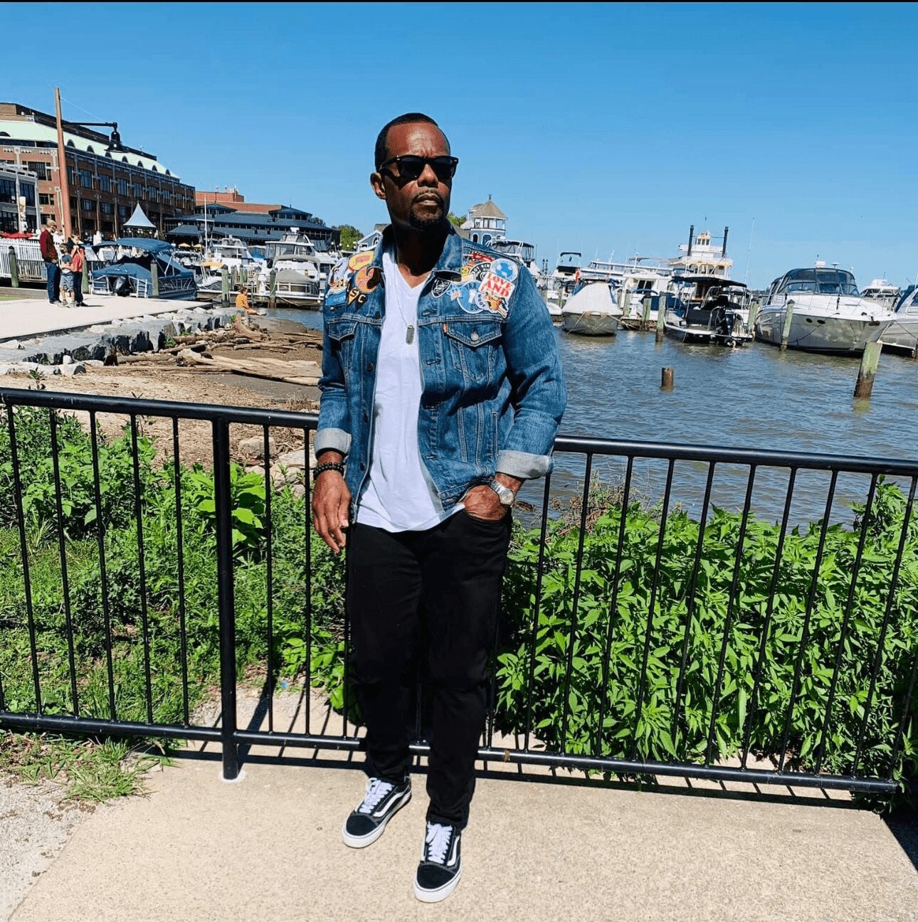 Man wearing sunglasses, denim jacket, white shirt, black pants, and sneakers standing by a black fence near water with boats and buildings in background.