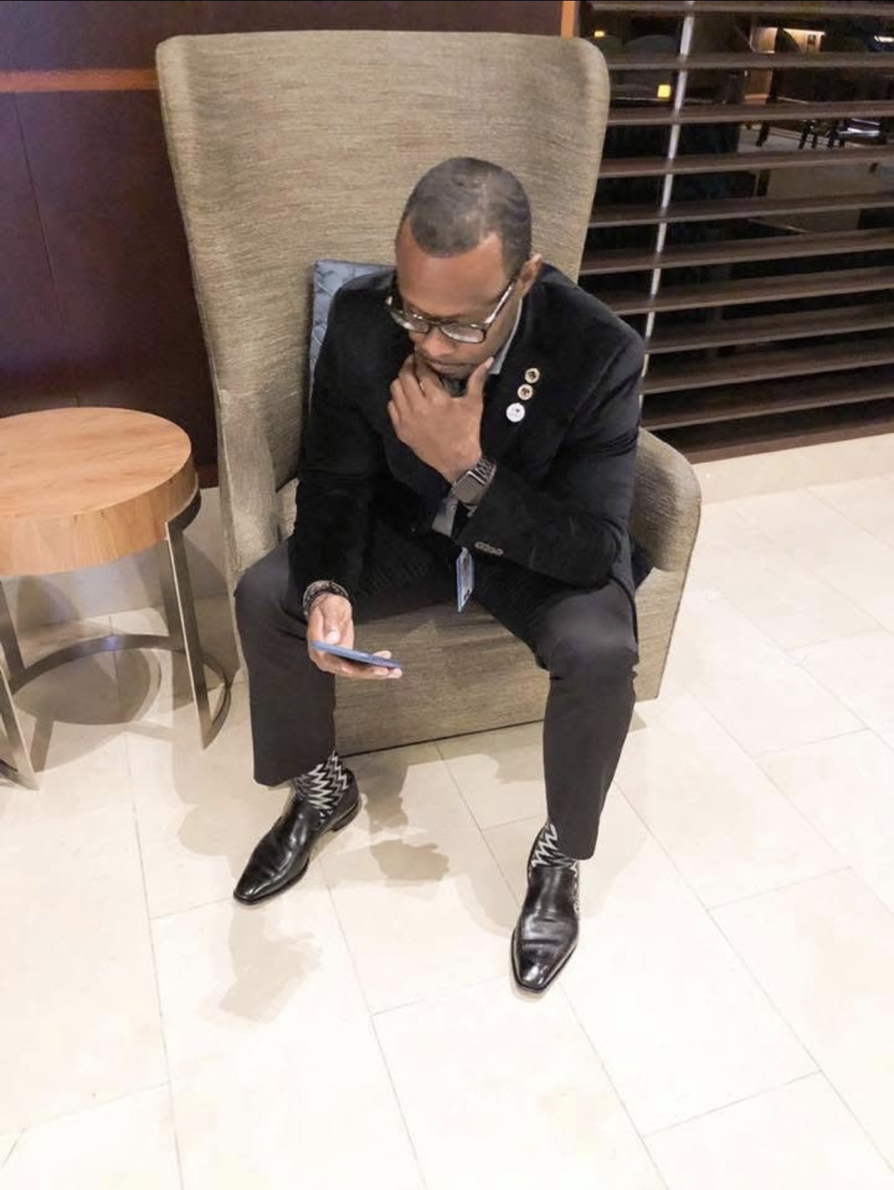 Person sitting in a large chair, looking at a phone, with a hand on chin, wearing glasses and formal attire.