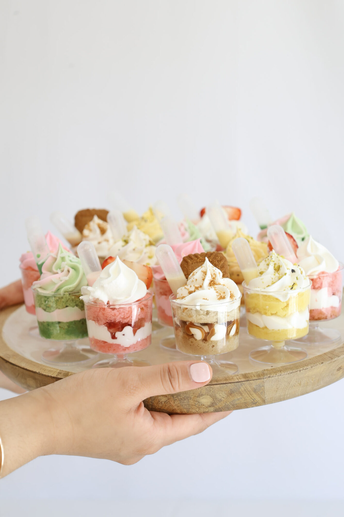 Hand holding a round wooden tray with colorful mini cakes and cupcakes, against a plain white background.