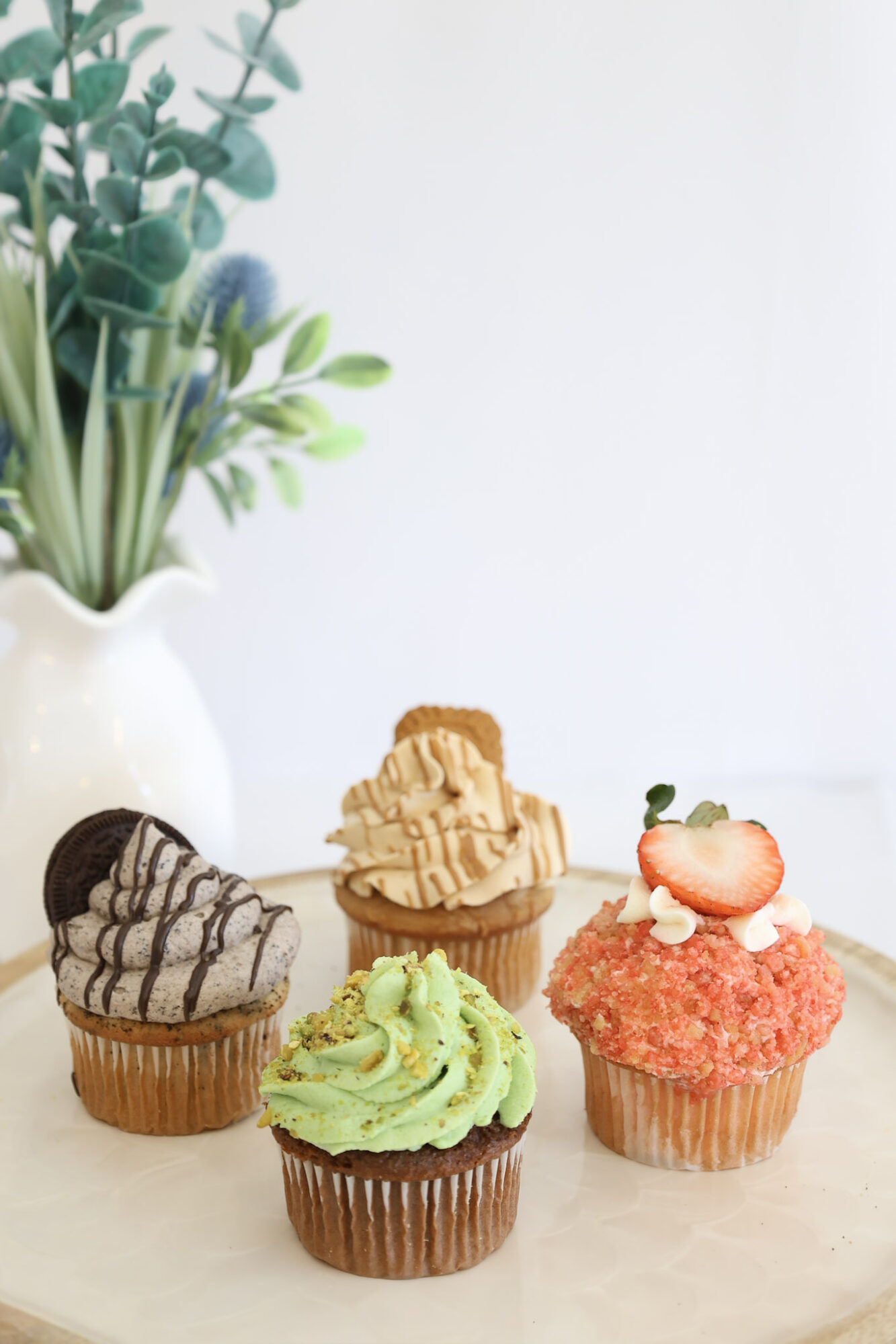 Five cupcakes with different toppings and colors arranged on a light surface, with a white vase and green plant in the background.