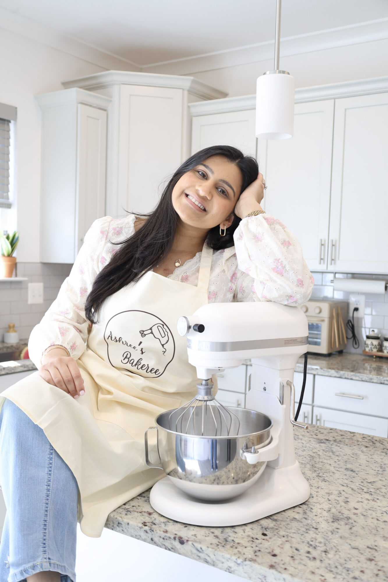 Woman in a kitchen wearing an apron, smiling, with a stand mixer on the counter, white cabinets in background.