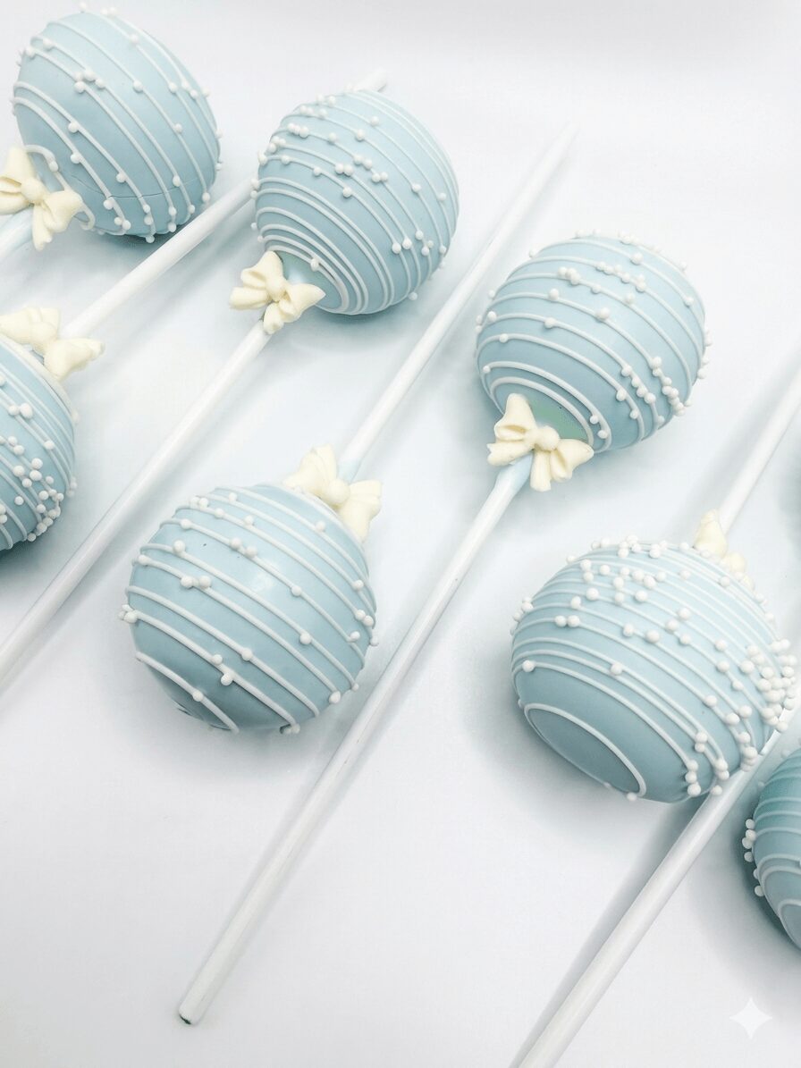Light blue cake pops with white drizzle and small white decorations, arranged on white sticks on a white surface.