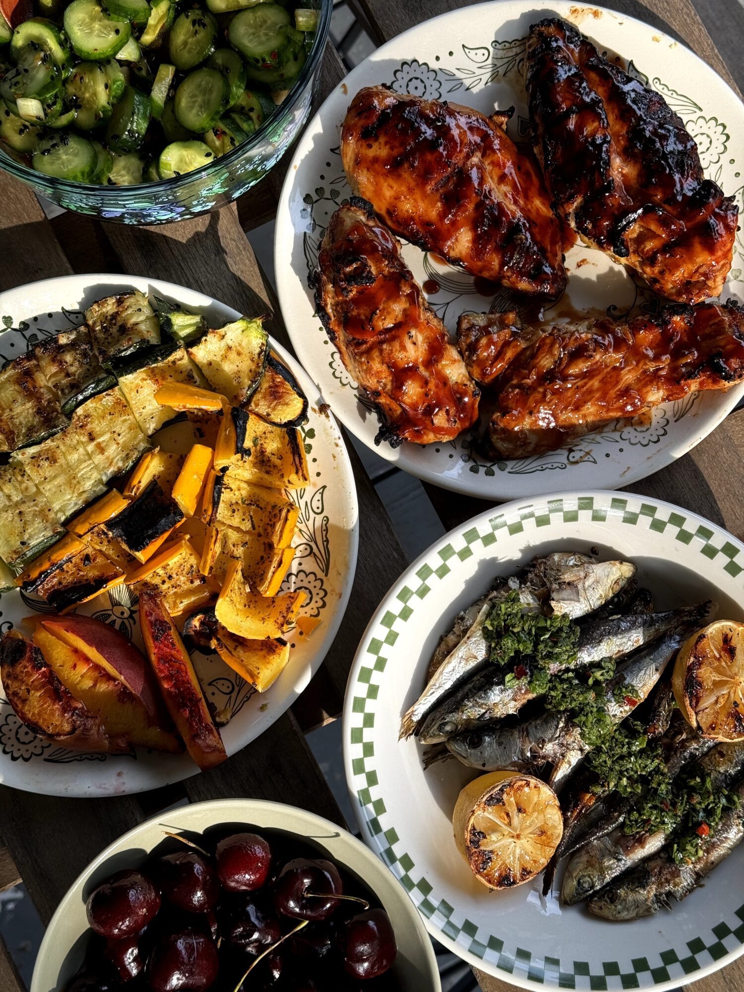 Plate of grilled chicken wings, grilled fish, grilled sardines, roasted eggplant, and a bowl of cherries.