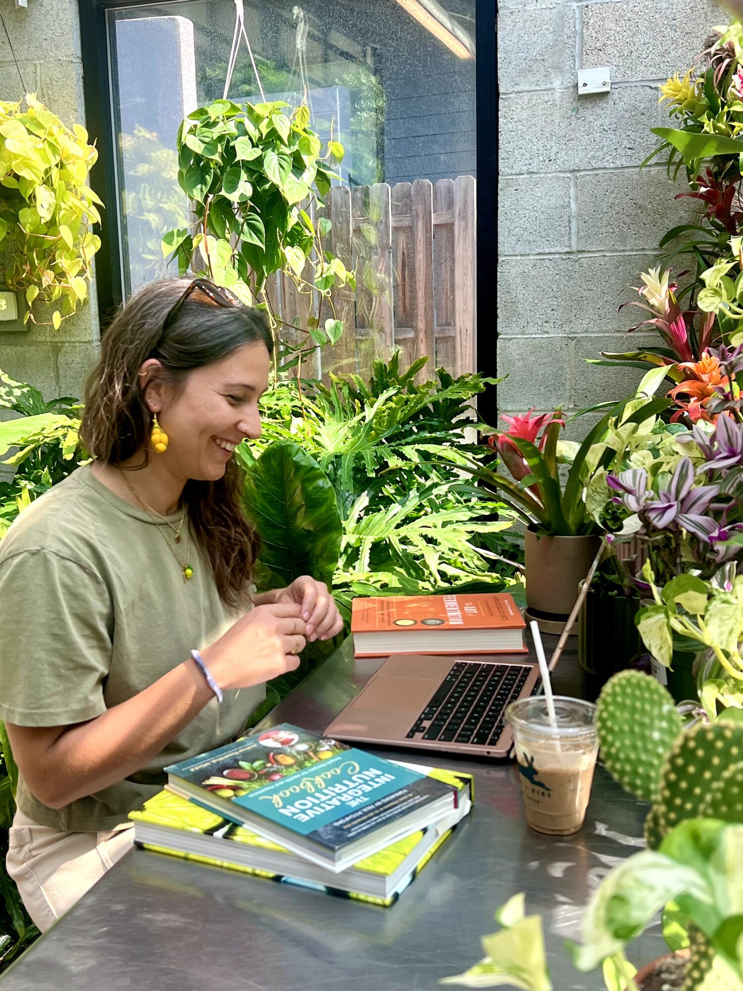 Woman sitting at a table outdoors with books, laptop, and plants, smiling and looking at her hands.