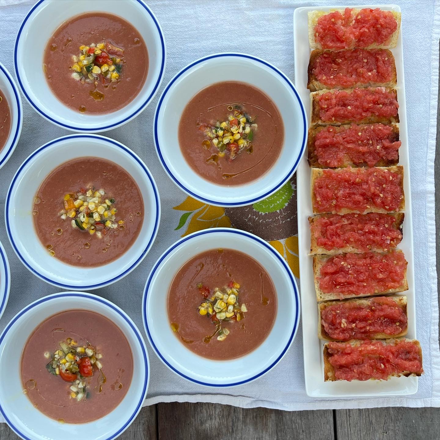 Five bowls of soup with toppings and a rectangular plate of sliced bread with red topping, on a tablecloth.
