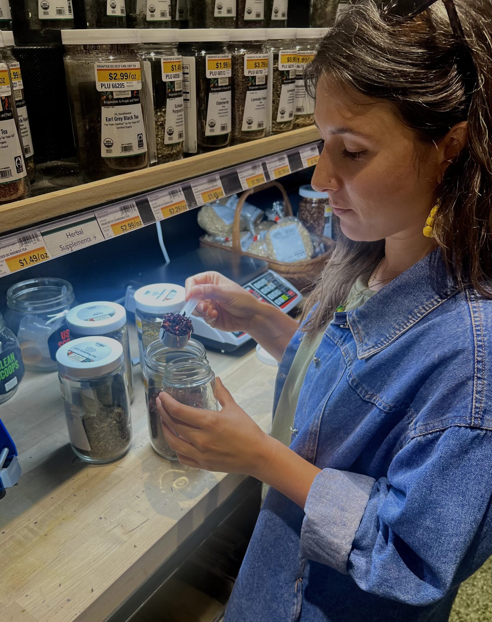 Young woman in denim jacket shopping for spices at grocery store, holding a jar and examining products on shelf.