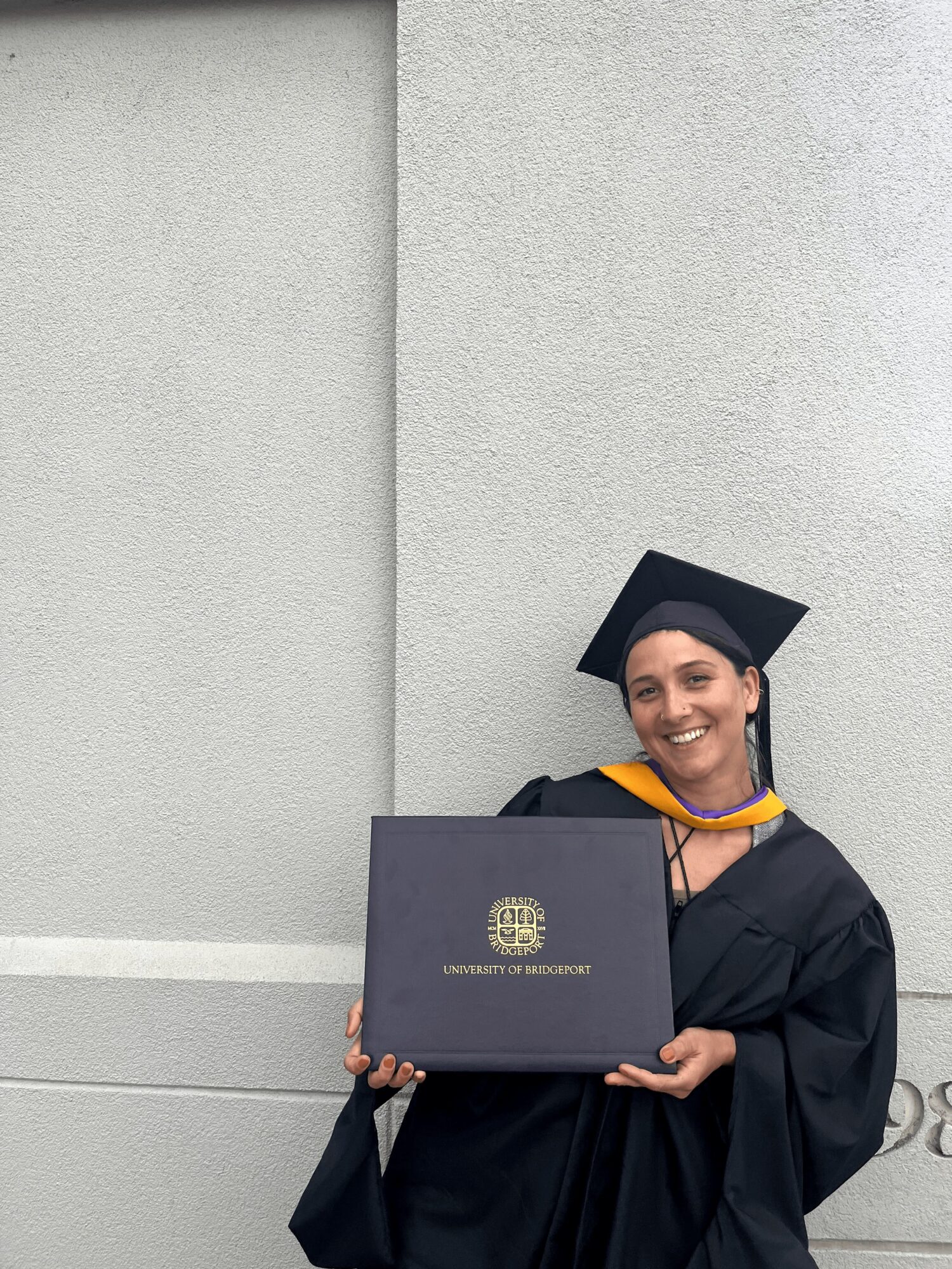 Graduate in cap and gown holding diploma in front of a plain wall, smiling.