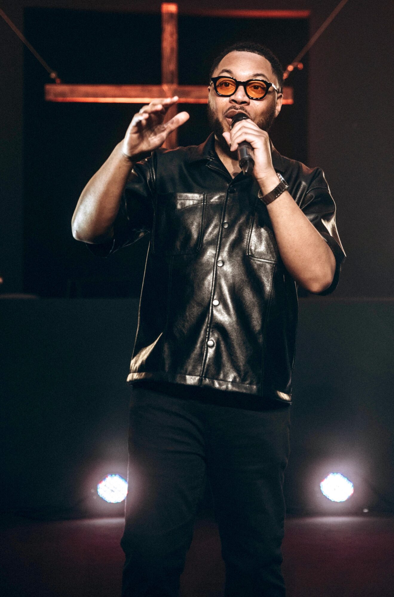Person speaking into microphone on stage, wearing glasses and a shiny black shirt, with stage lights behind.
