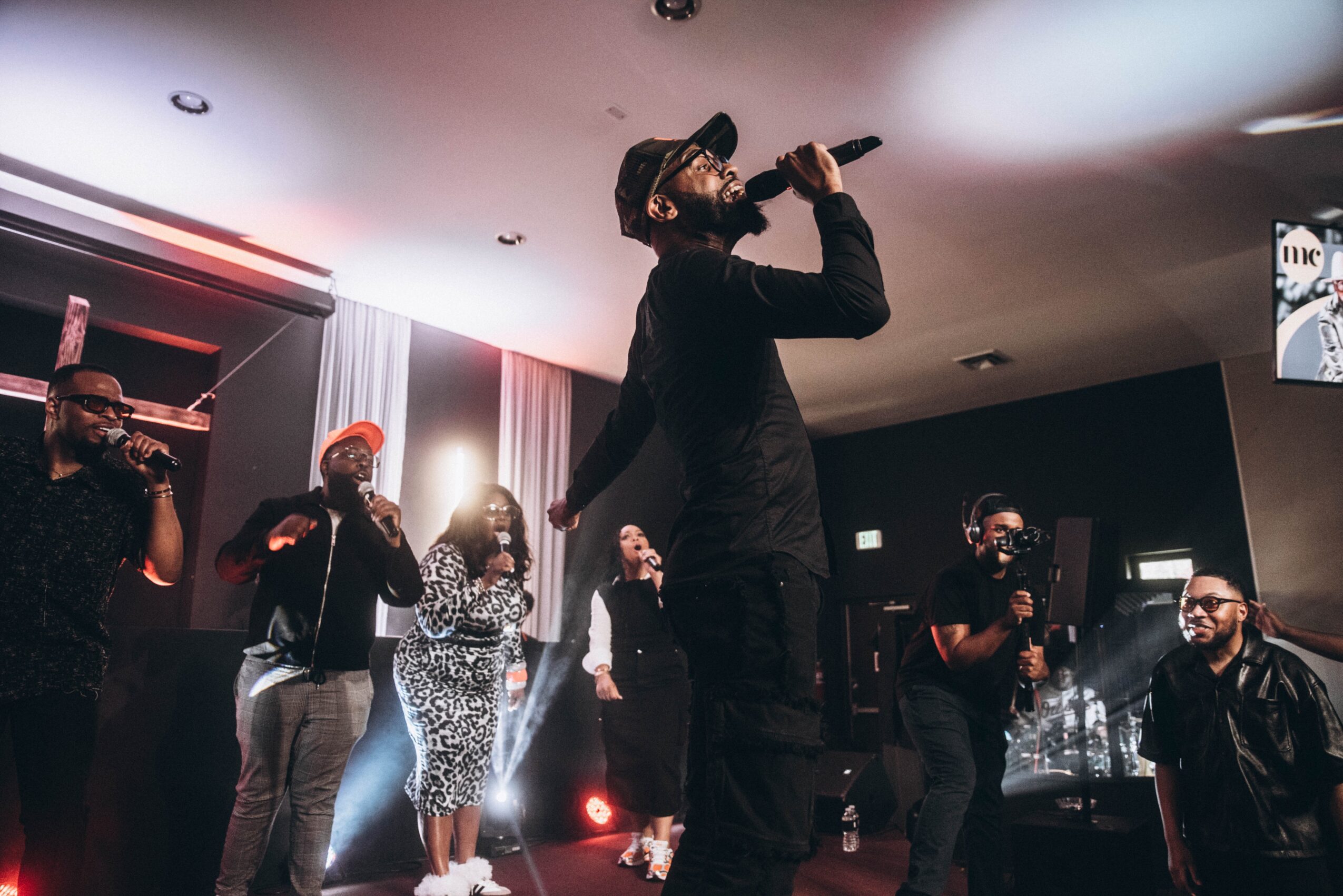 A man in black clothing and a hat performs with a microphone on stage, surrounded by five people, some with microphones, in a dimly lit room.