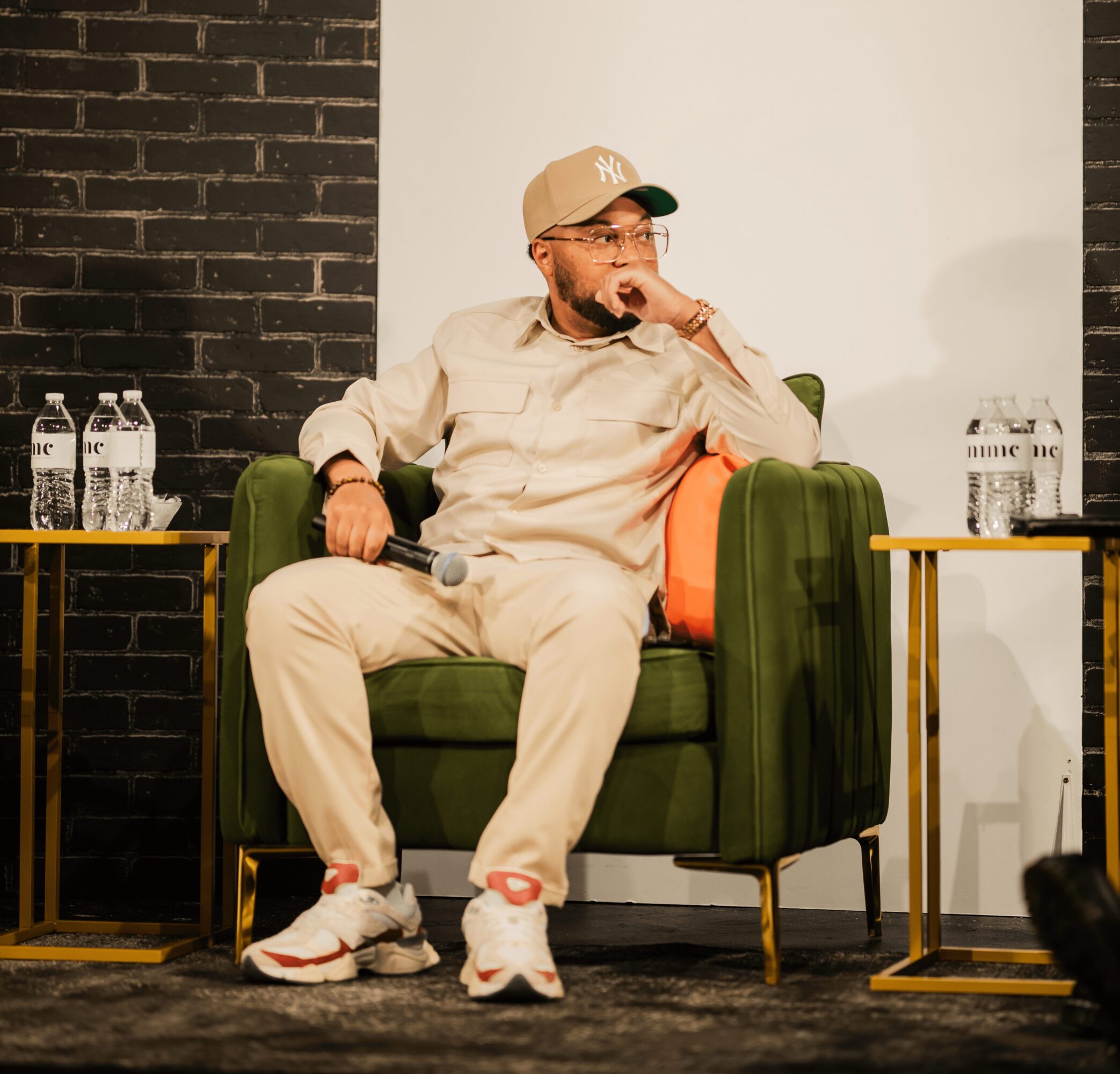 Person sitting on a green armchair, wearing a beige cap and outfit, holding a microphone, with two tables and water bottles nearby.