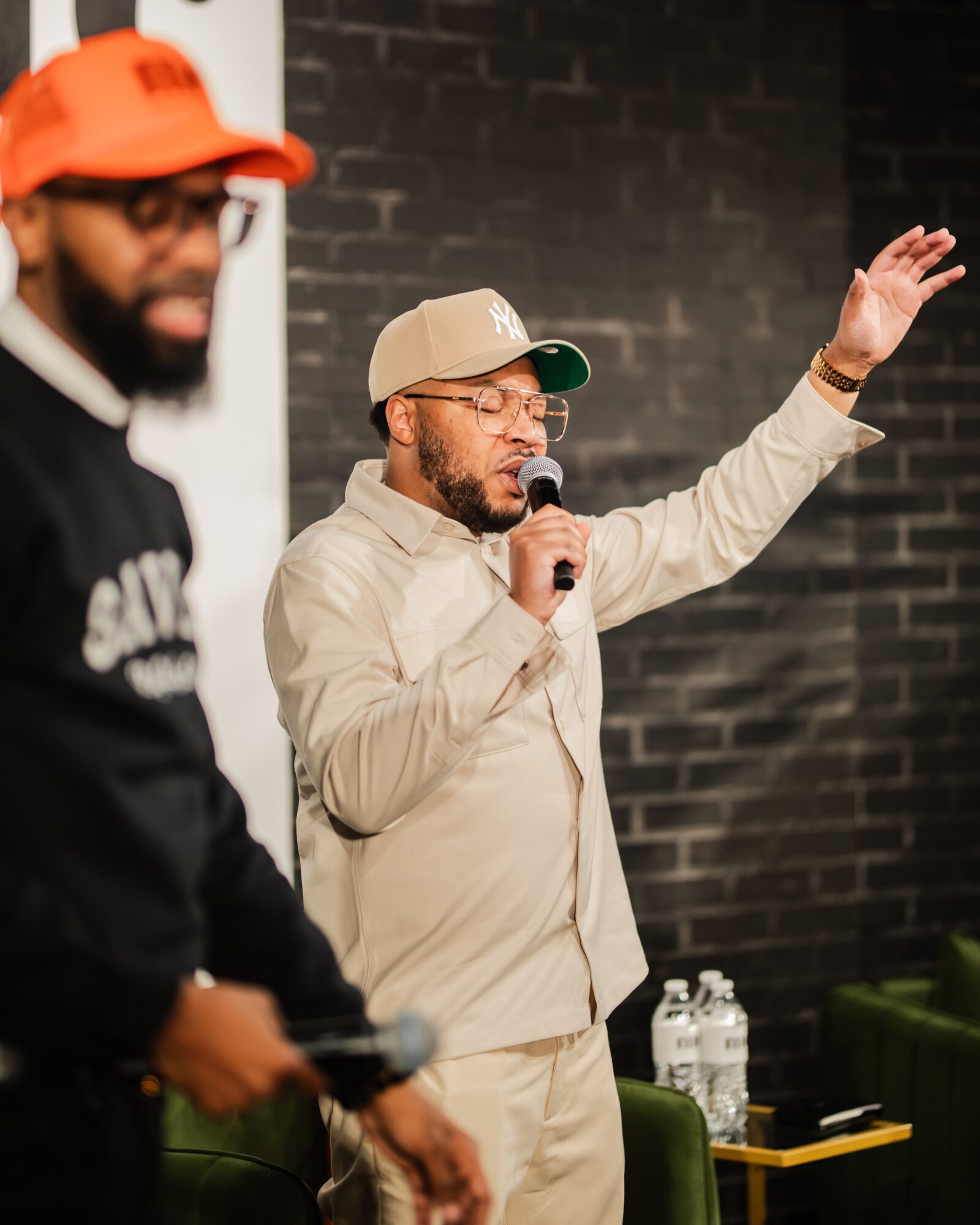 Man speaking into microphone with raised hand, wearing beige jacket and cap, in front of dark brick wall.