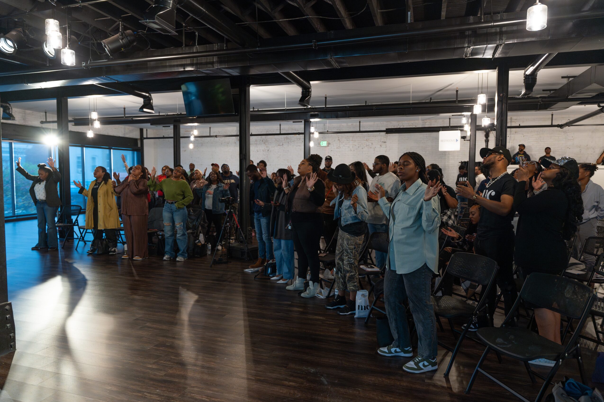 Group of people standing in a room with large windows, some with hands raised, facing a speaker or presentation.