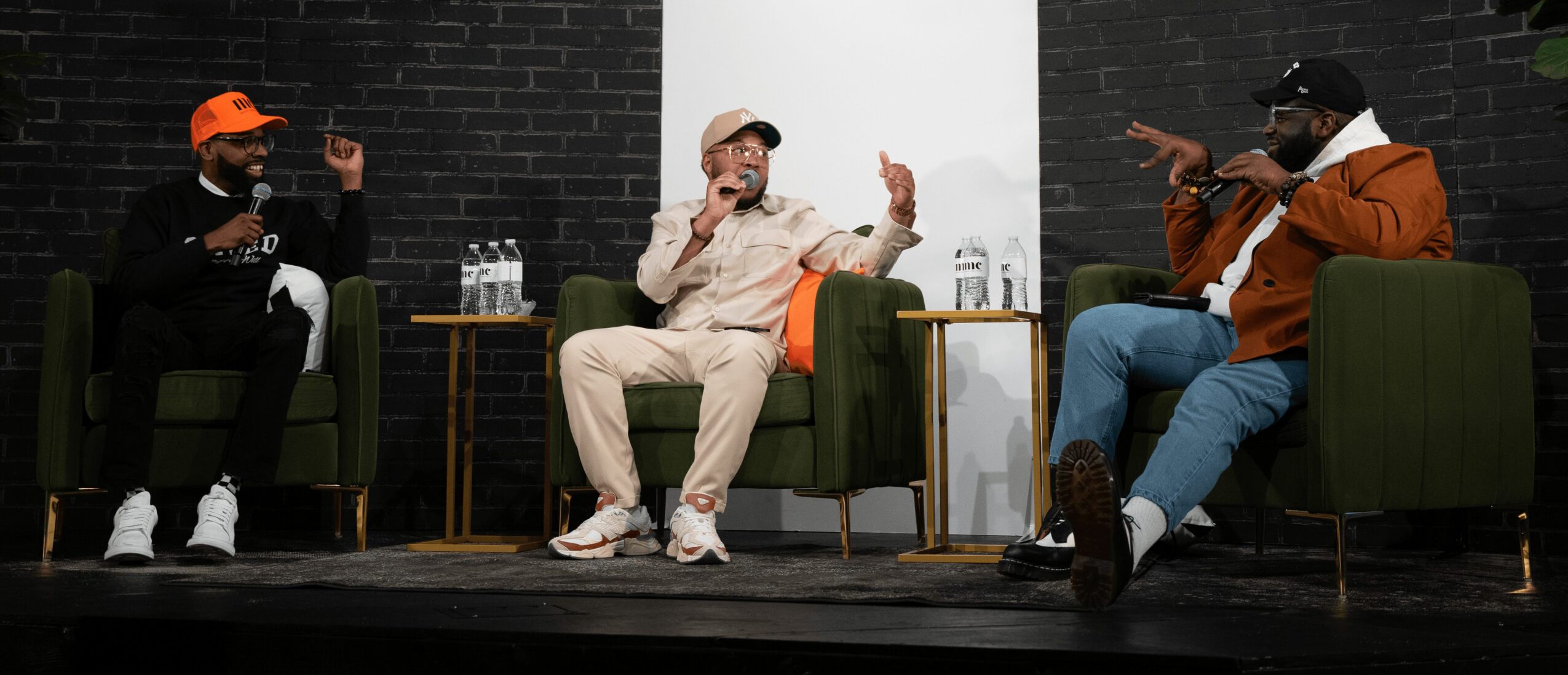 Three people sitting on green chairs in a room with black walls, engaged in conversation, with a white panel behind them.