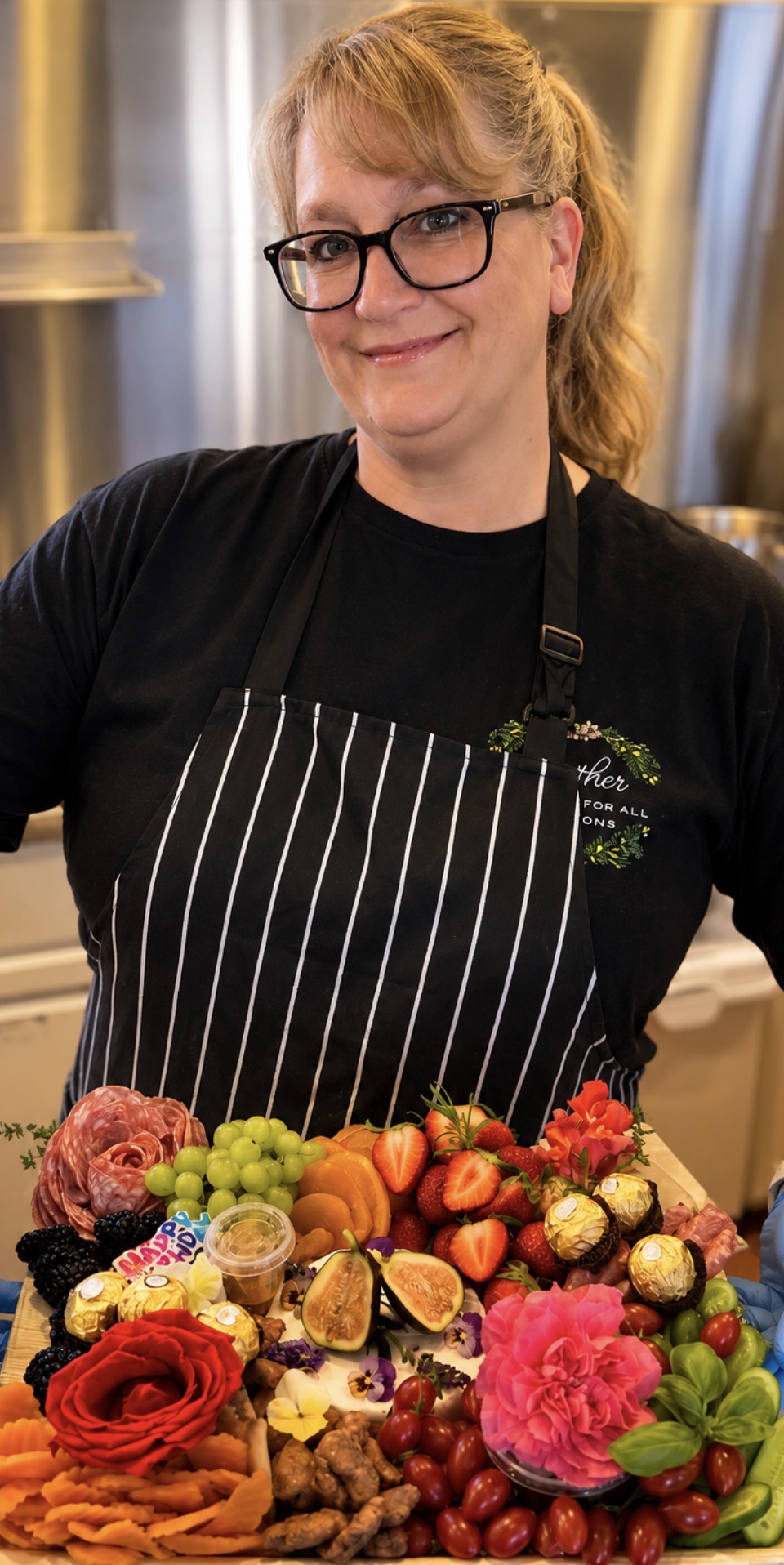 Woman with glasses and apron standing behind a colorful display of flowers and fruits in a kitchen.