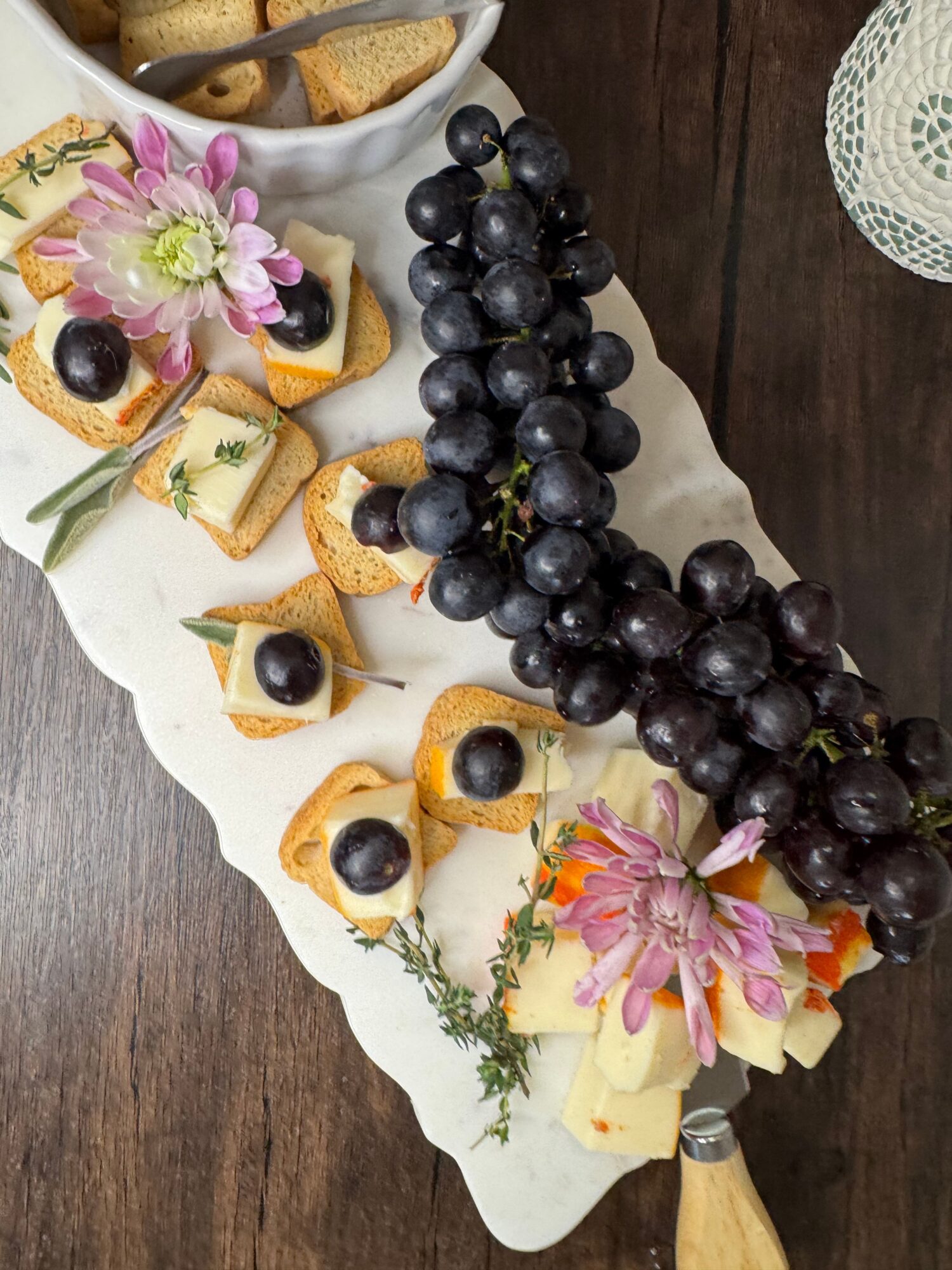 A white platter with grapes, cheese, crackers, and pink flowers on a dark wooden surface.