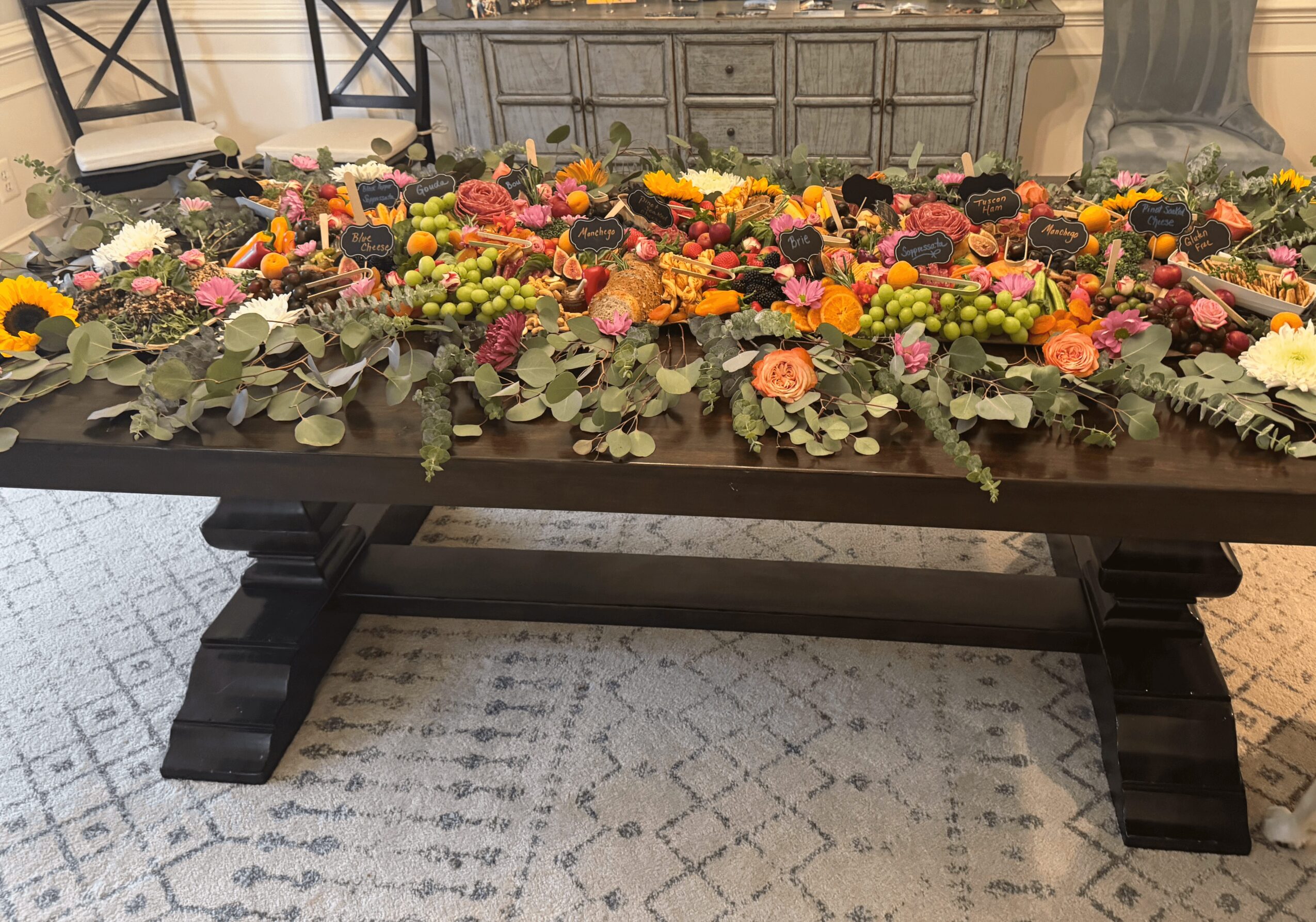 Table with colorful flowers and foliage, set in a room with chairs and a cabinet in the background.