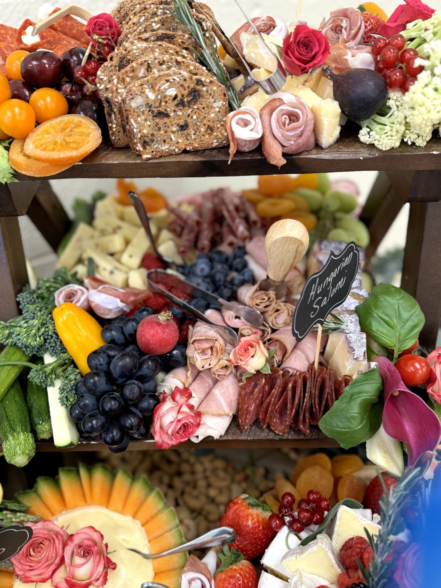Three-tiered fruit and cheese display with grapes, berries, cheese, flowers, and a chalkboard sign, arranged on a dark table.